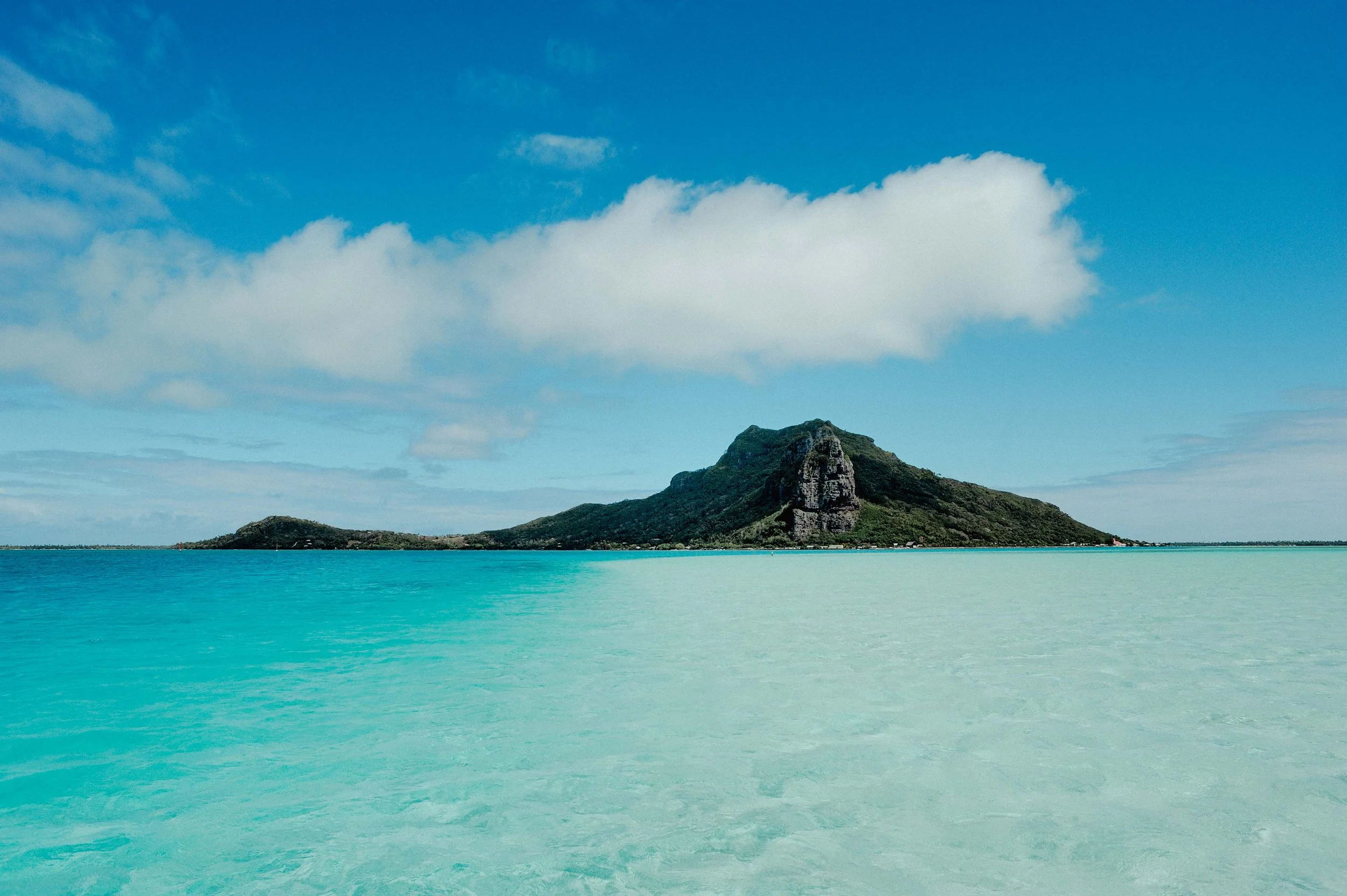Une île avec une montagne au centre, entourée d'une mer turquoise claire et un ciel bleu avec quelques nuages.