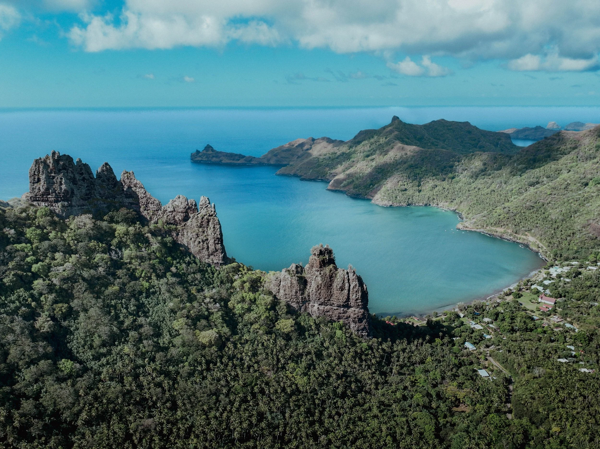 Vue d'une baie entourée de montagnes verdoyantes et d'une forêt dense avec des formations rocheuses, sur fond d'océan et ciel bleu avec quelques nuages.