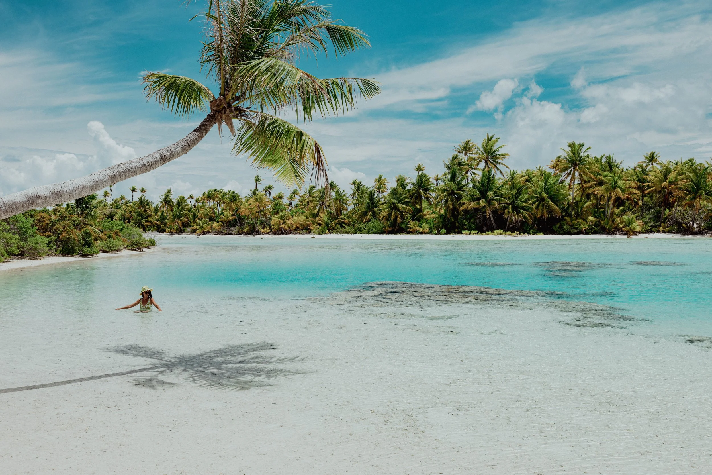 Plage de sable blanc avec un arbre de cocotier penché, eau turquoise calme, forêt dense de cocotiers en arrière-plan, ciel bleu avec quelques nuages, une personne portant un chapeau de soleil nage dans la mer.