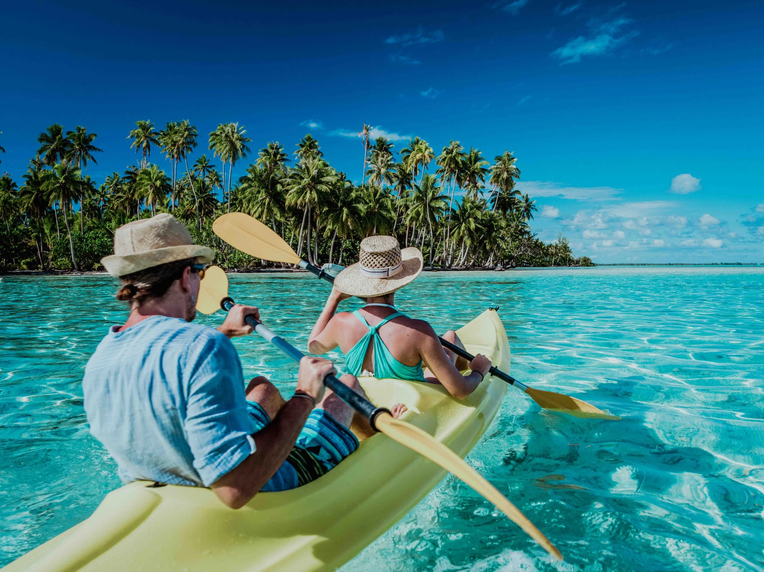 Deux personnes pagayant en kayak dans une eau turquoise, avec une végétation de palmiers en arrière-plan, sous un ciel bleu avec quelques nuages.