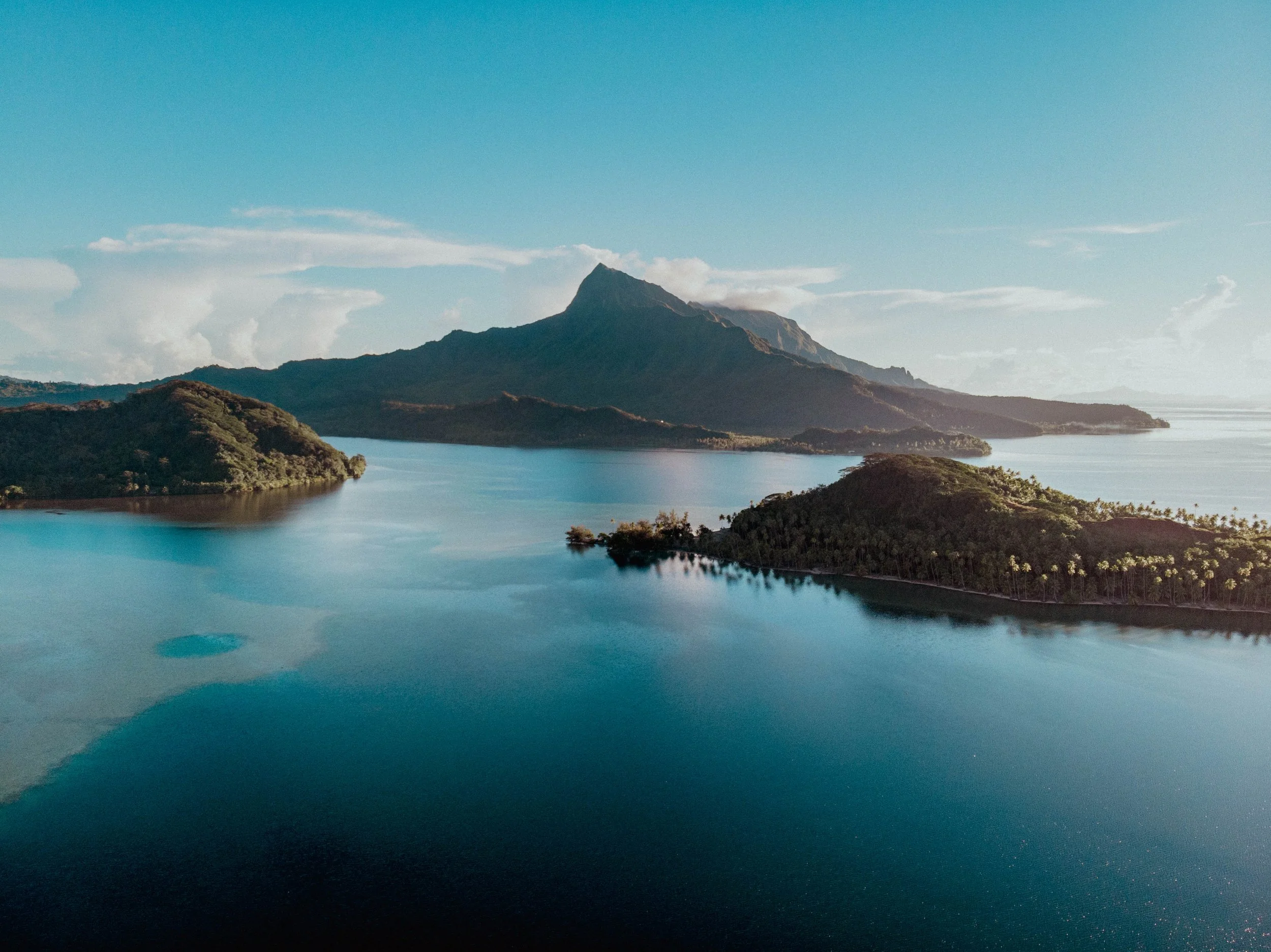 Une vue aérienne d'une grande étendue d'eau entourée de collines verdoyantes, avec une montagne en arrière-plan sous un ciel bleu clair avec quelques nuages.
