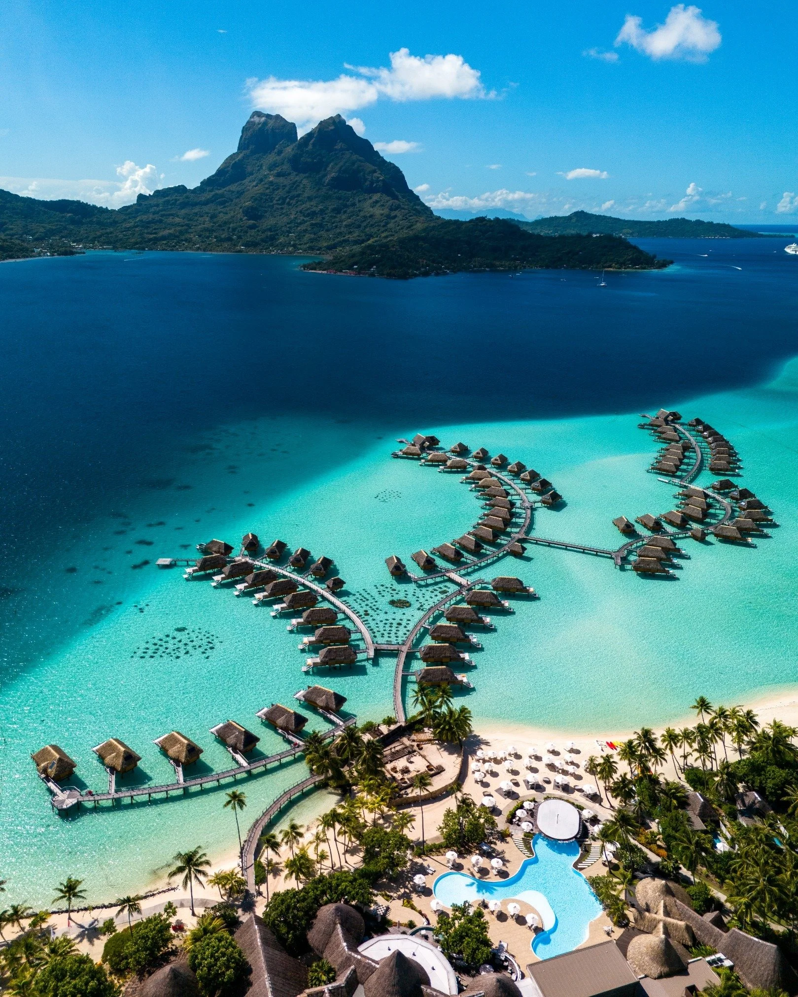 vue aérienne d'une station balnéaire avec des bungalows sur pilotis au-dessus de l'eau turquoise, entourée de palmiers, avec une montagne en arrière-plan et un ciel bleu.