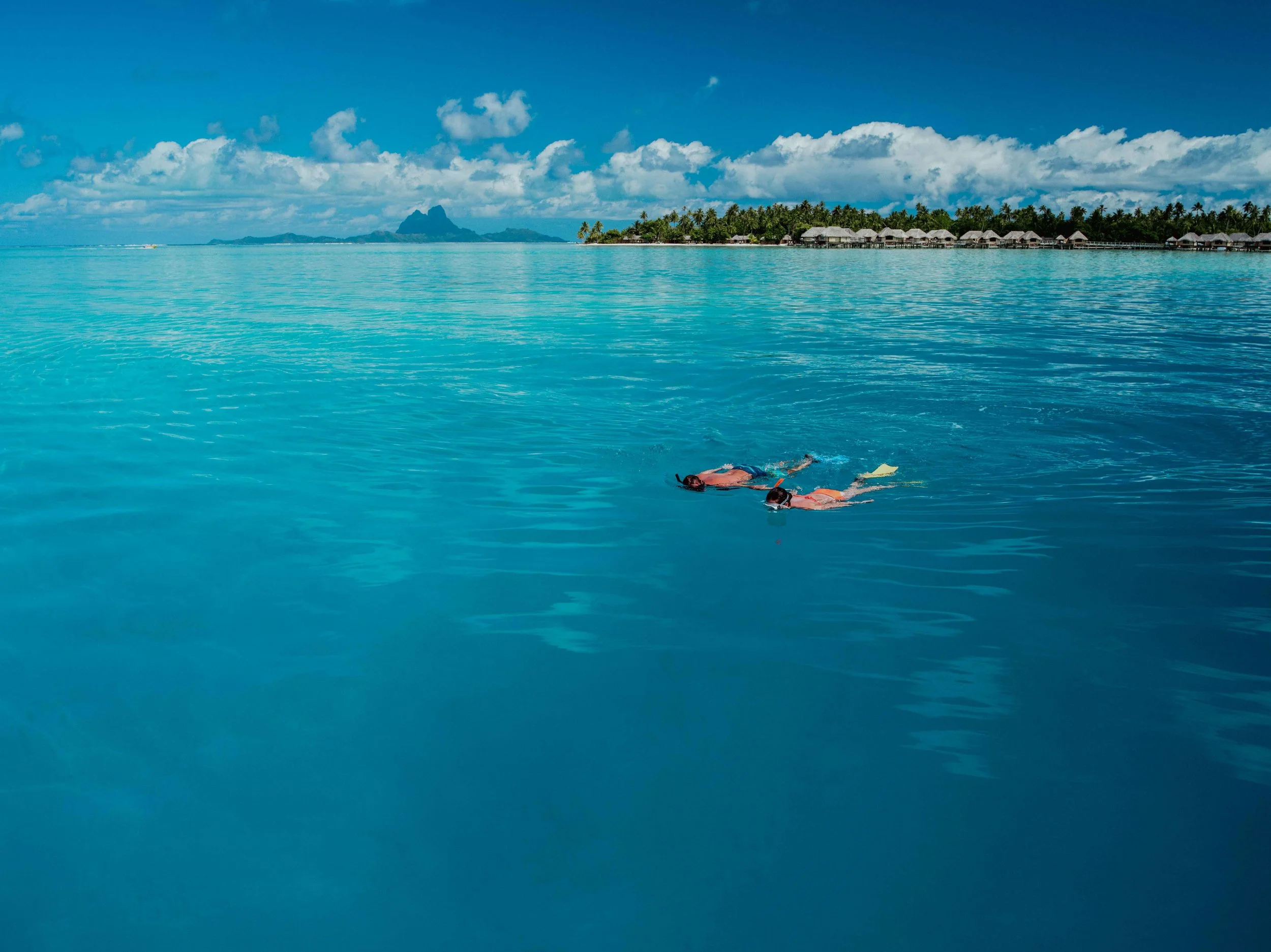 Deux personnes nageant dans une mer turquoise avec une île et des overwater bavilions en arrière-plan sous un ciel partiellement nuageux.