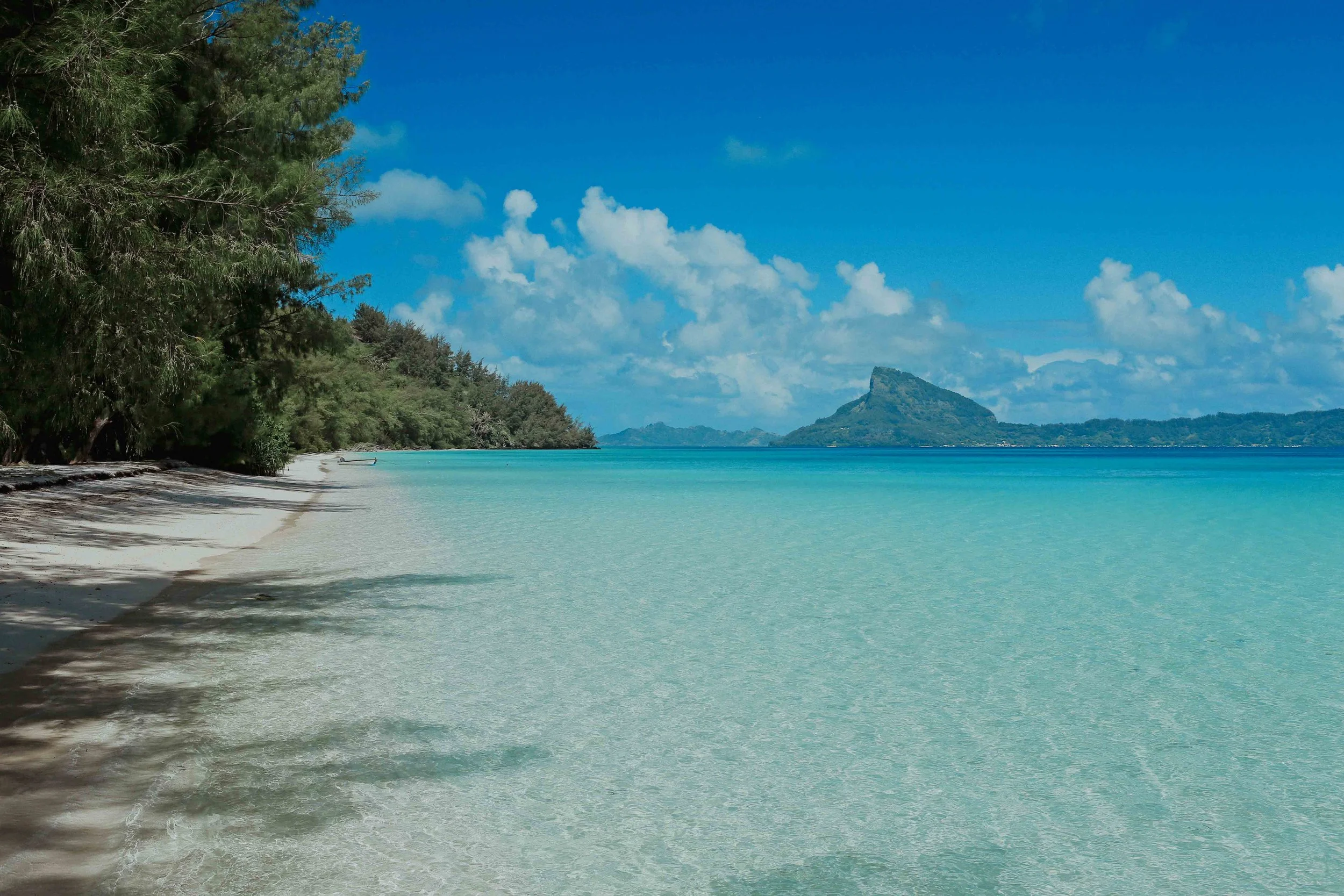 Plage de sable blanc avec des eaux turquoise, bordée d'arbres verts, avec une montagne en forme distincte à l'horizon et un ciel bleu avec quelques nuages.