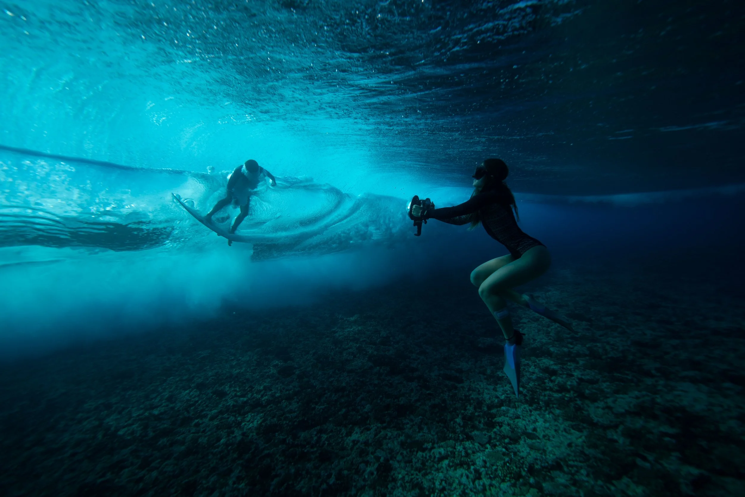 Une femme en combinaison de plongée et nageoires documente un surfeur à l'intérieur d'une vague sous-marine avec une caméra.