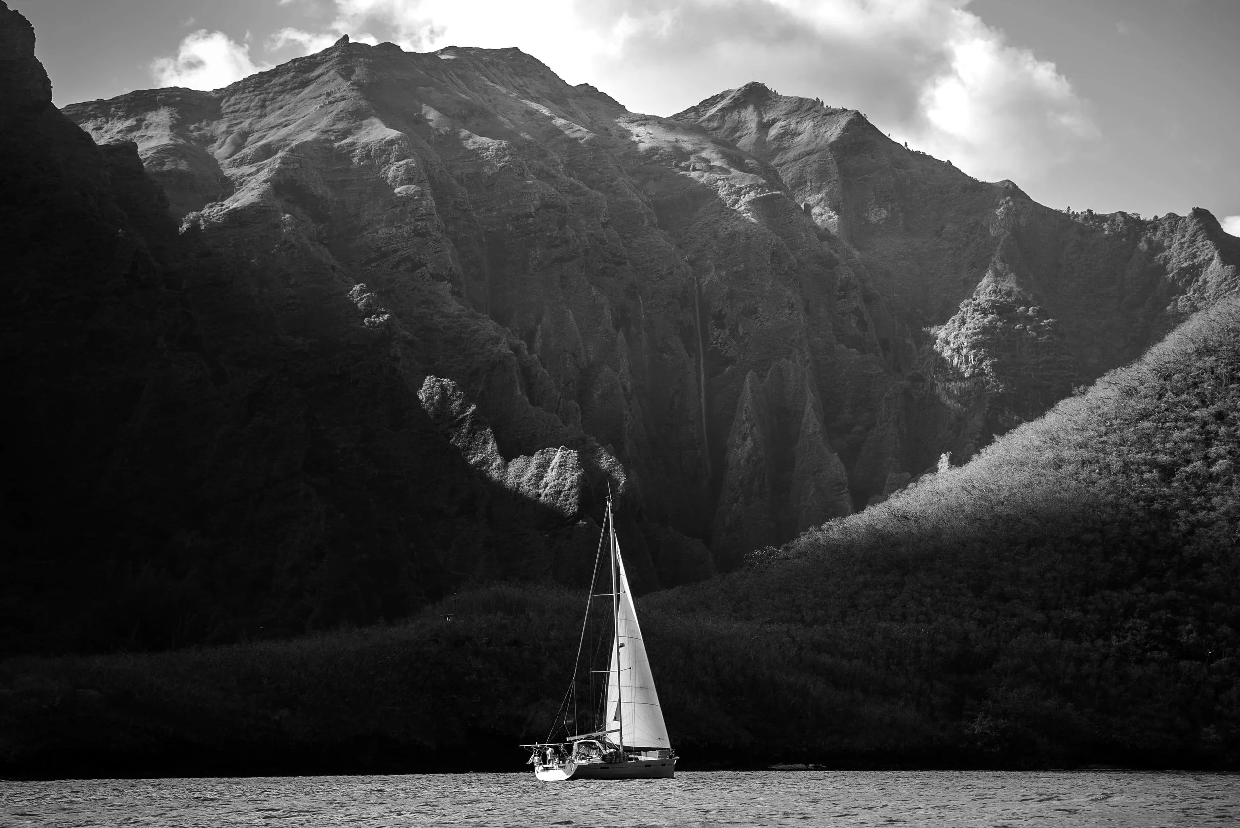 Un voilier naviguant sur une rivière entourée de montagnes escarpées et boisées dans un paysage en noir et blanc.