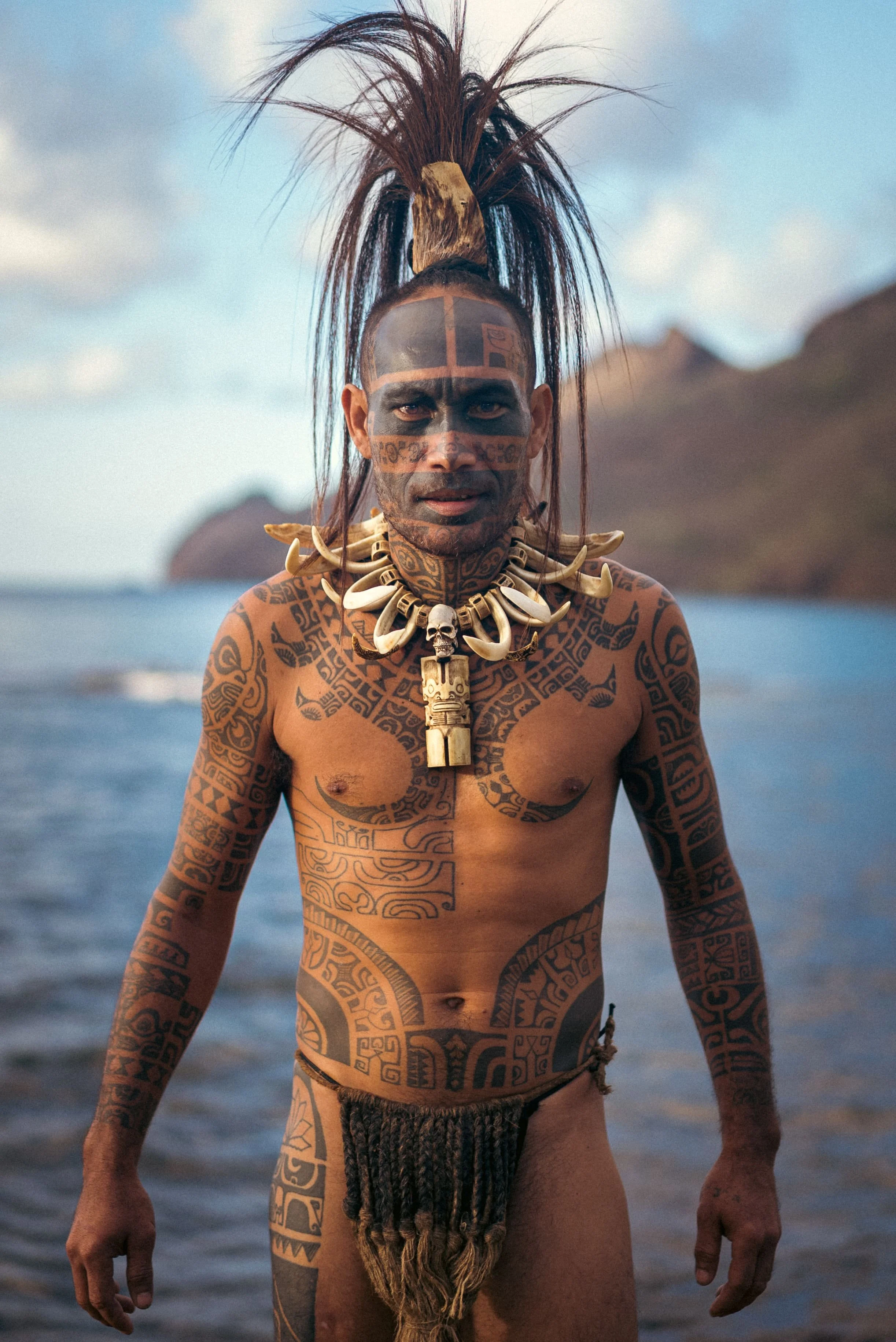 Un homme avec des tatouages traditionnels et des accessoires en ivoire, portant une coiffure élaborée, debout devant la mer avec un paysage montagneux en arrière-plan.