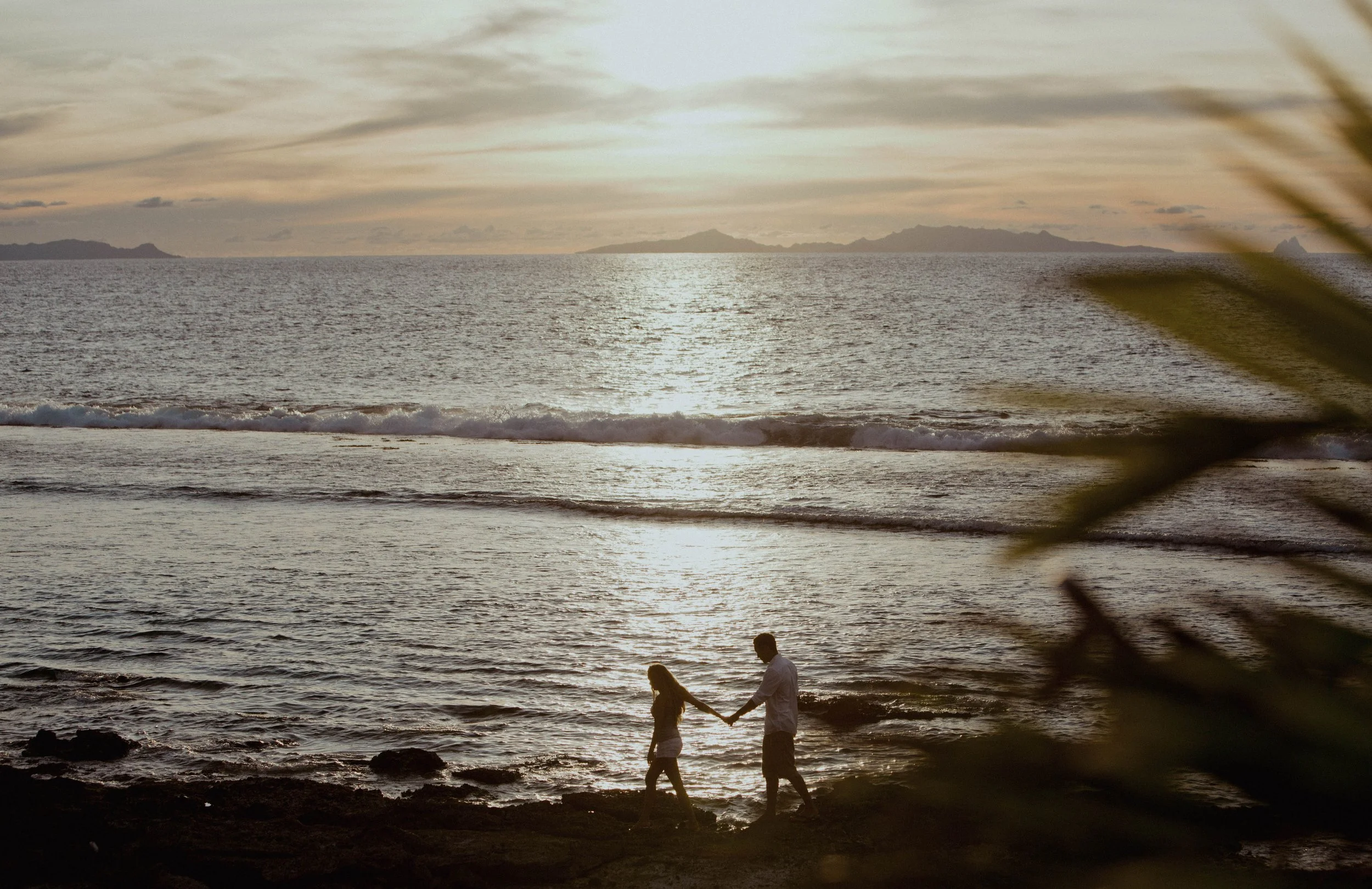 Un couple qui marche sur la plage au coucher du soleil, avec des montagnes à l'horizon et des feuilles floues au premier plan.