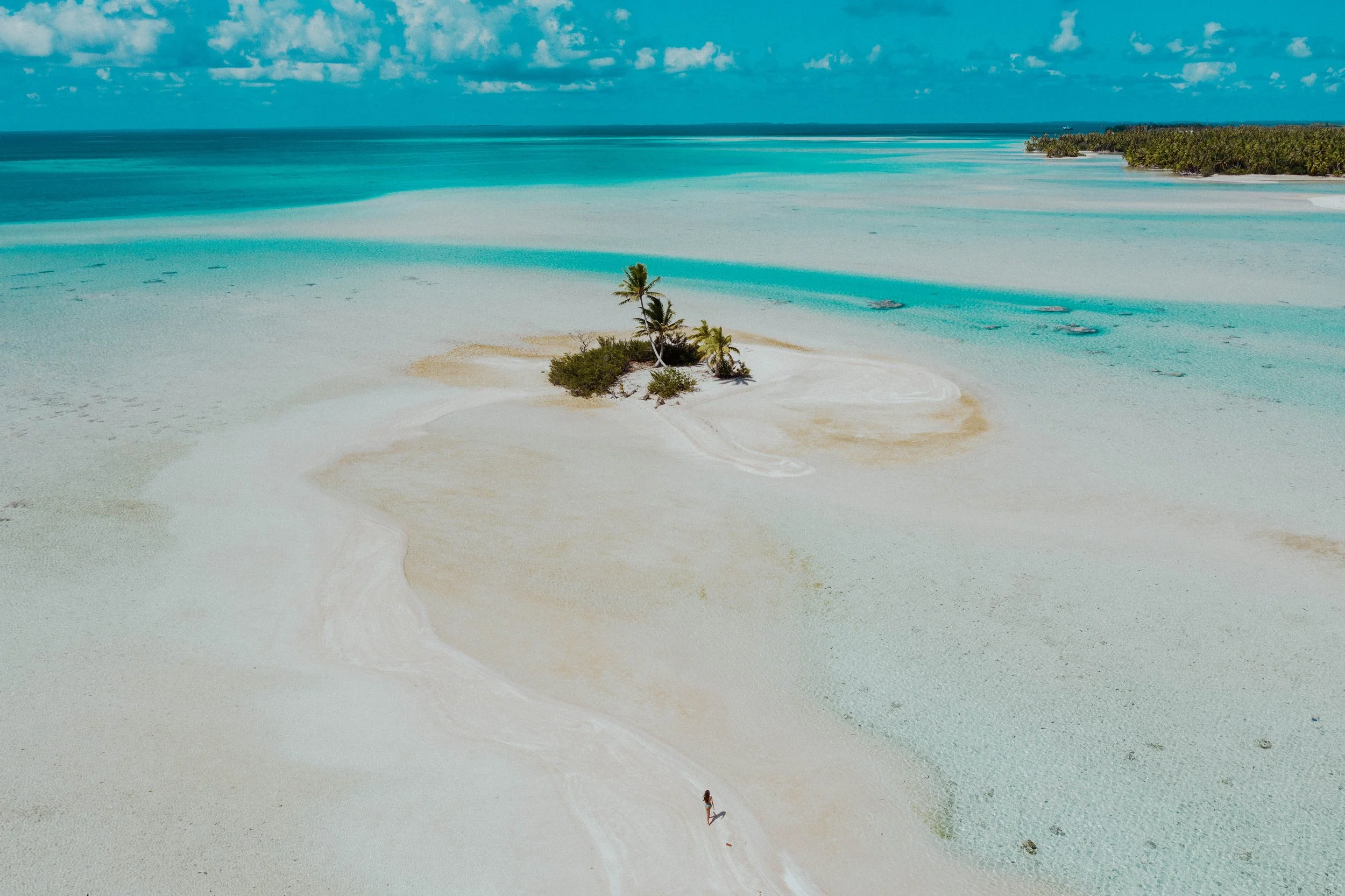 Plage de sable blanc avec un petit arbre, vue aérienne, eaux turquoise, ciel bleu avec quelques nuages.