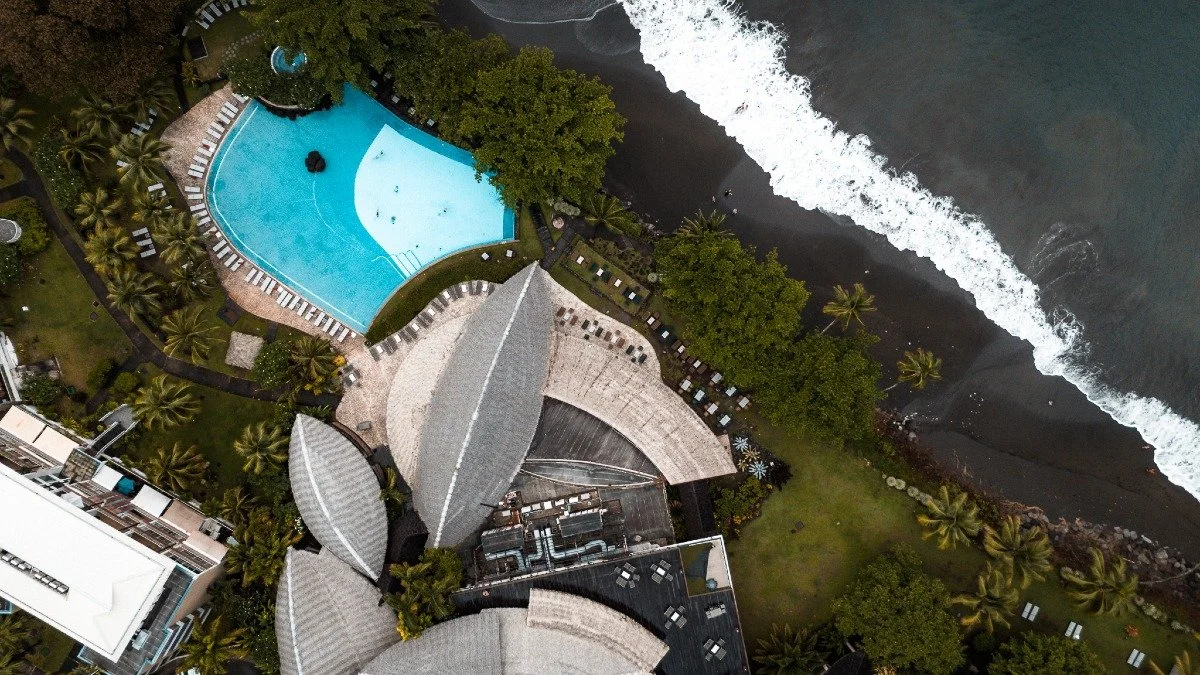 Vue aérienne d'une piscine en forme de cerf-volant, d'une plage de sable noir, et de bâtiments entourés de palmiers, vue sur l'océan