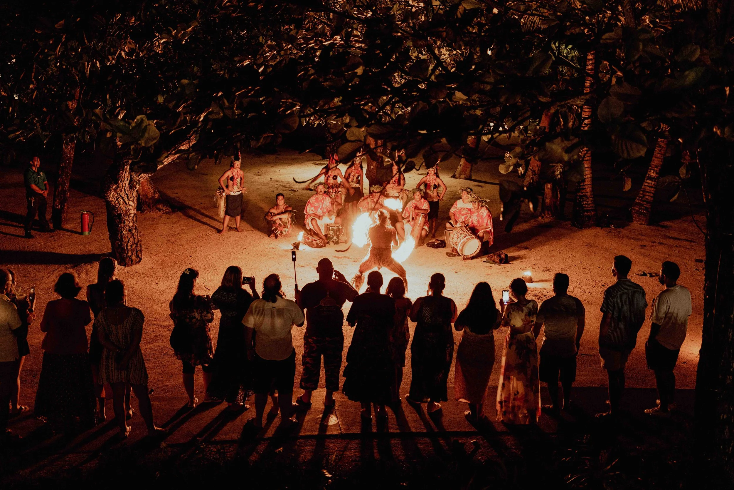 Un groupe de personnes assises autour d'un feu de camp, regardant une performance de guerriers avec des torches, dans une scène nocturne en plein air. Des arbres entourent la scène.