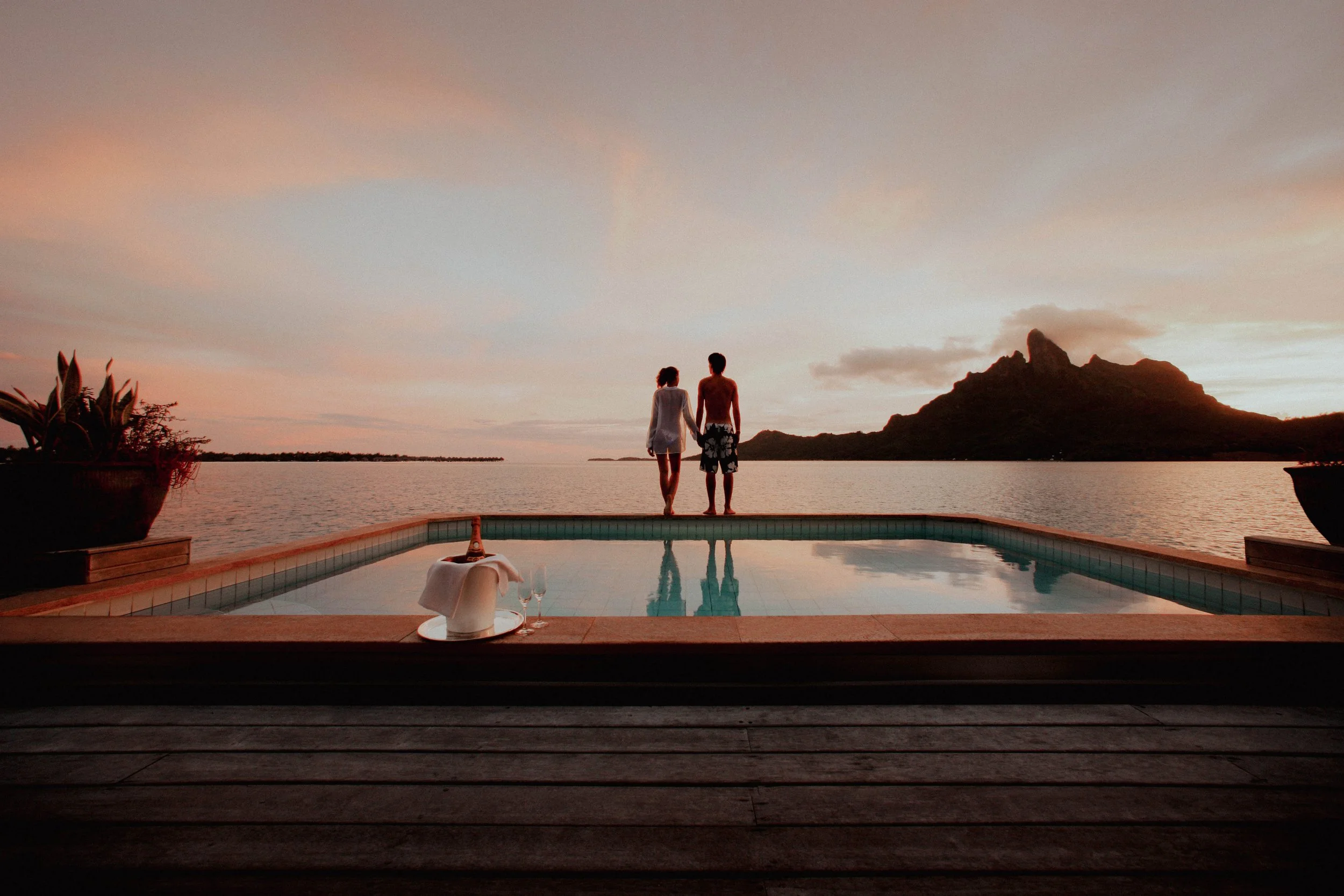 Un couple debout au bord d'une piscine regardant le coucher de soleil sur un lac et une montagne.