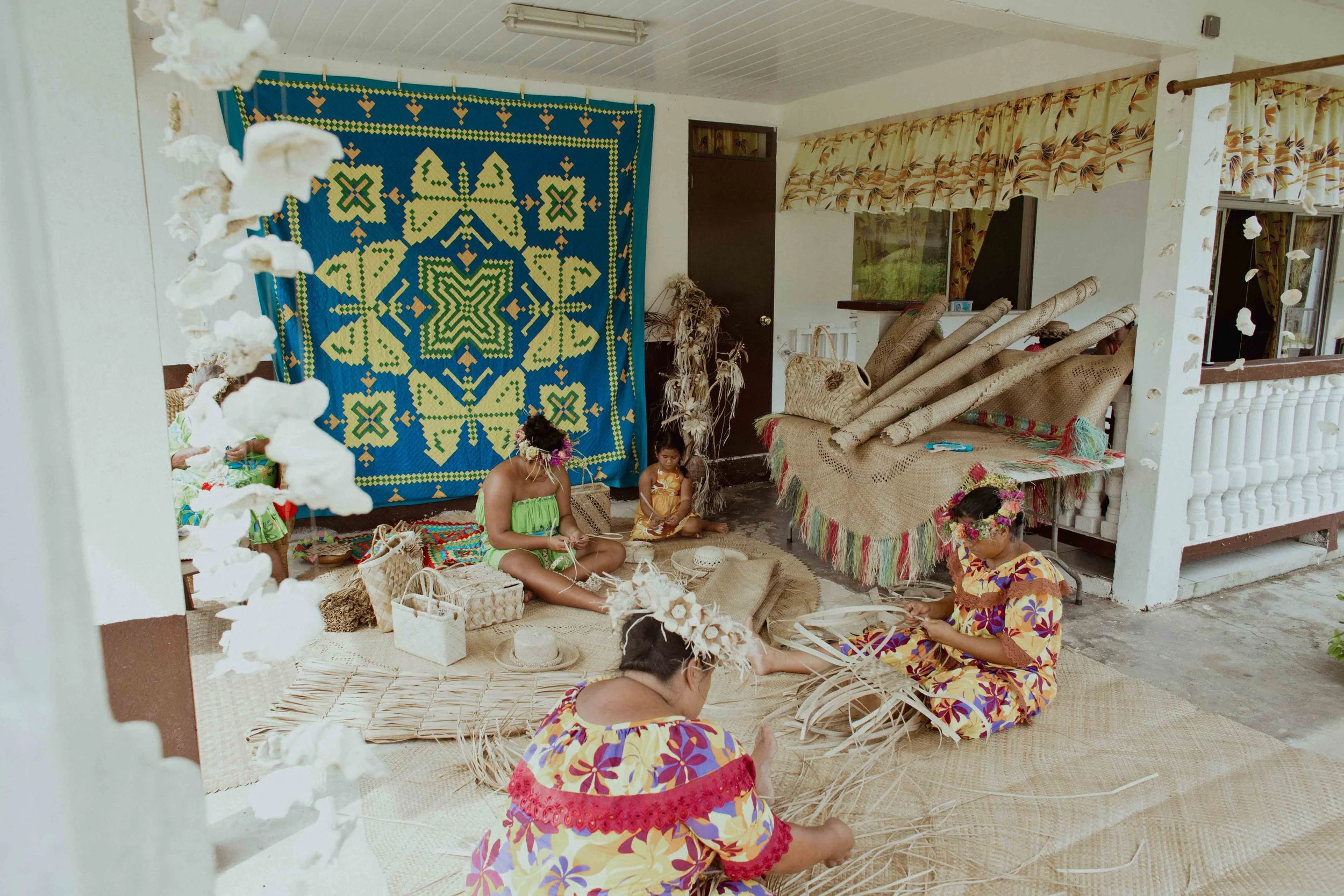 Groupe de femmes et d'enfants en vêtements traditionnels colorés, travaillant avec des matériaux naturels comme des feuilles et des fibres dans une pièce décorée de tapis et de textiles colorés, avec un mur en tissu brodé en arrière-plan.