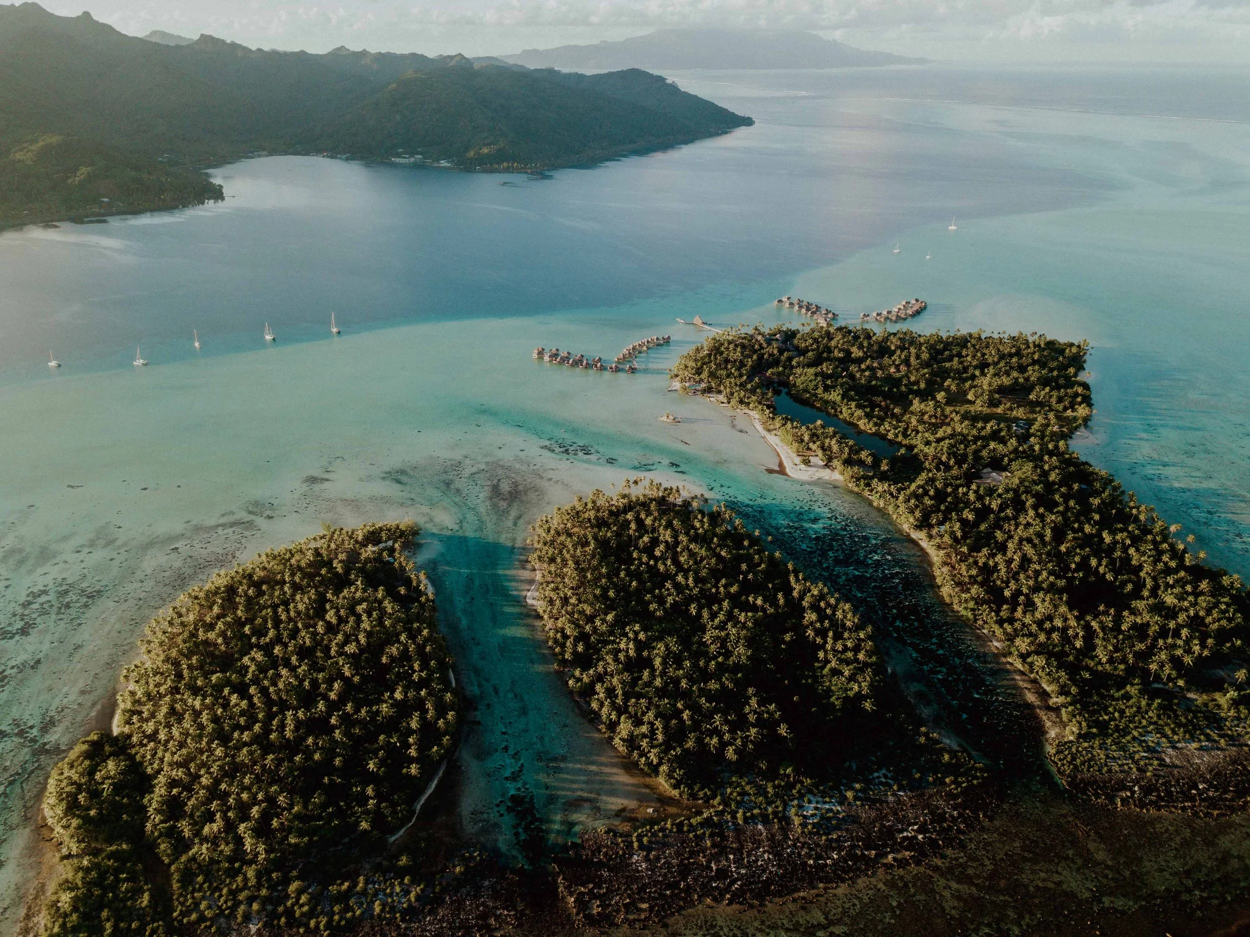 Paysage aérien d'une île tropicale couverte de végétation dense, entourée d'eau turquoise avec quelques yachts. Des bungalows sur pilotis sont visibles au large de l'île.