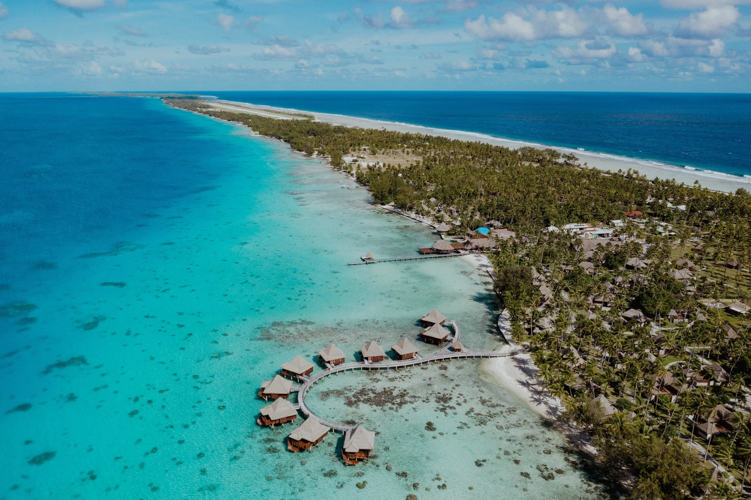 Vue aérienne d'une plage tropicale avec des bungalows sur pilotis et un fin rivage en sable blanc entouré d'eau turquoise claire.