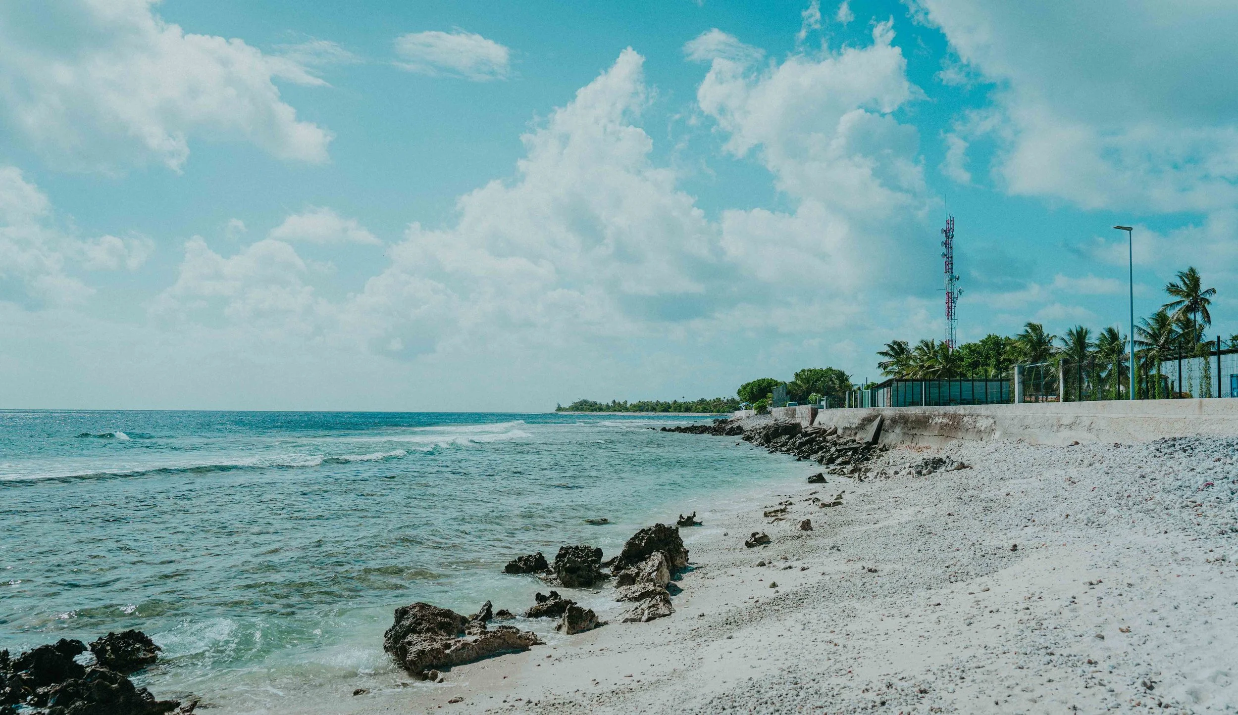 Plage avec sable clair, rochers noirs, mer calme, ciel bleu avec nuages, palmiers et bâtiments en arrière-plan.
