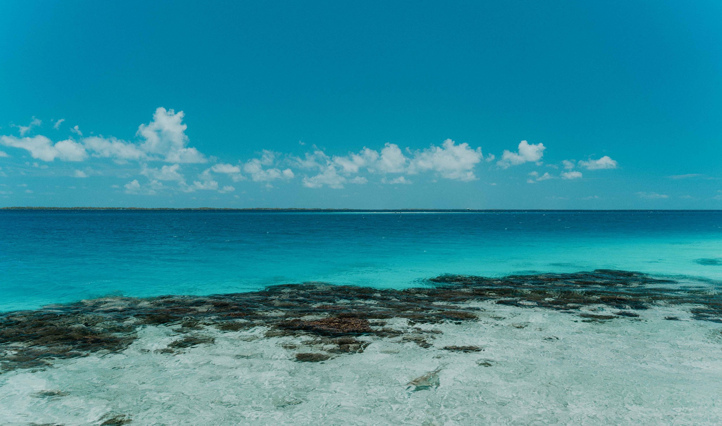 Plage de sable blanc avec rochers, eau turquoise et ciel bleu avec quelques nuages.