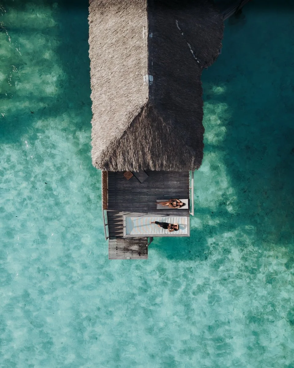 Deux femmes posent devant une cabane en bois construite sur l'eau turquoise d'une mer tropicale. La cabane a un toit en chaume et un pont en bois reliant la cabane à la plage.