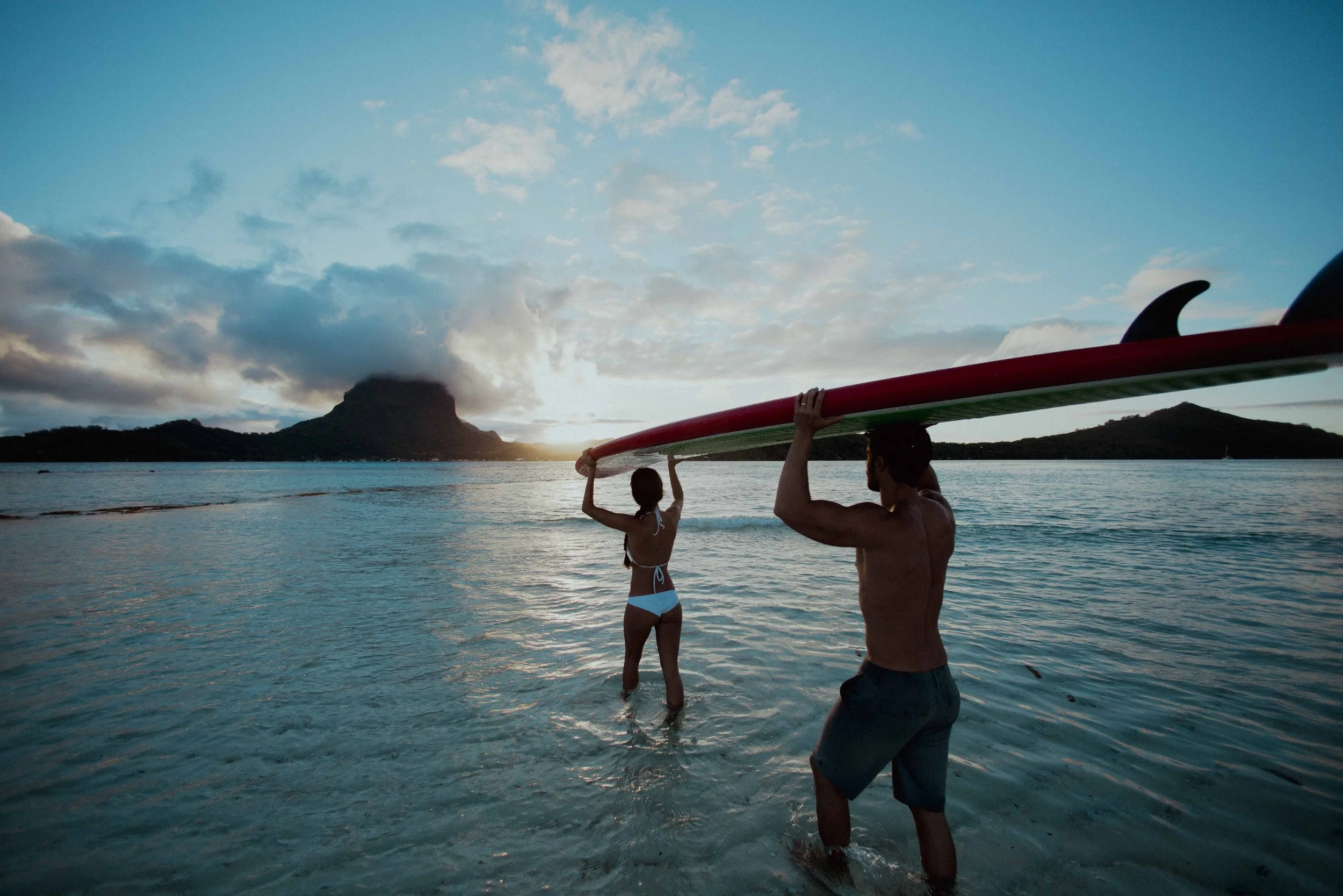 Un homme et une jeune fille avec un paddle surf sur une plage, au coucher du soleil, avec des montagnes en arrière-plan.
