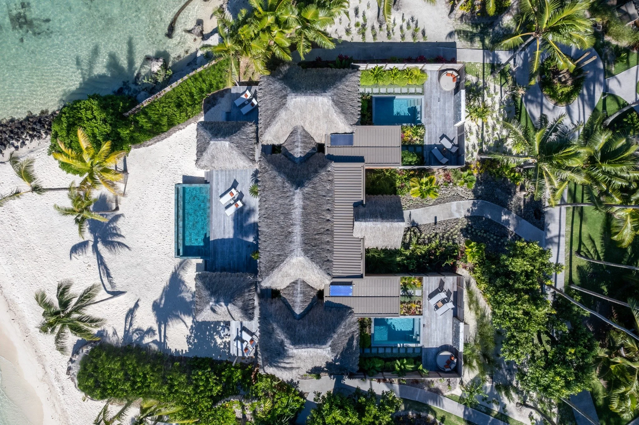 Plan aérien d'un complexe de vacances avec plusieurs villas, piscines, zones de détente, palmiers et une plage de sable blanc visible à gauche.