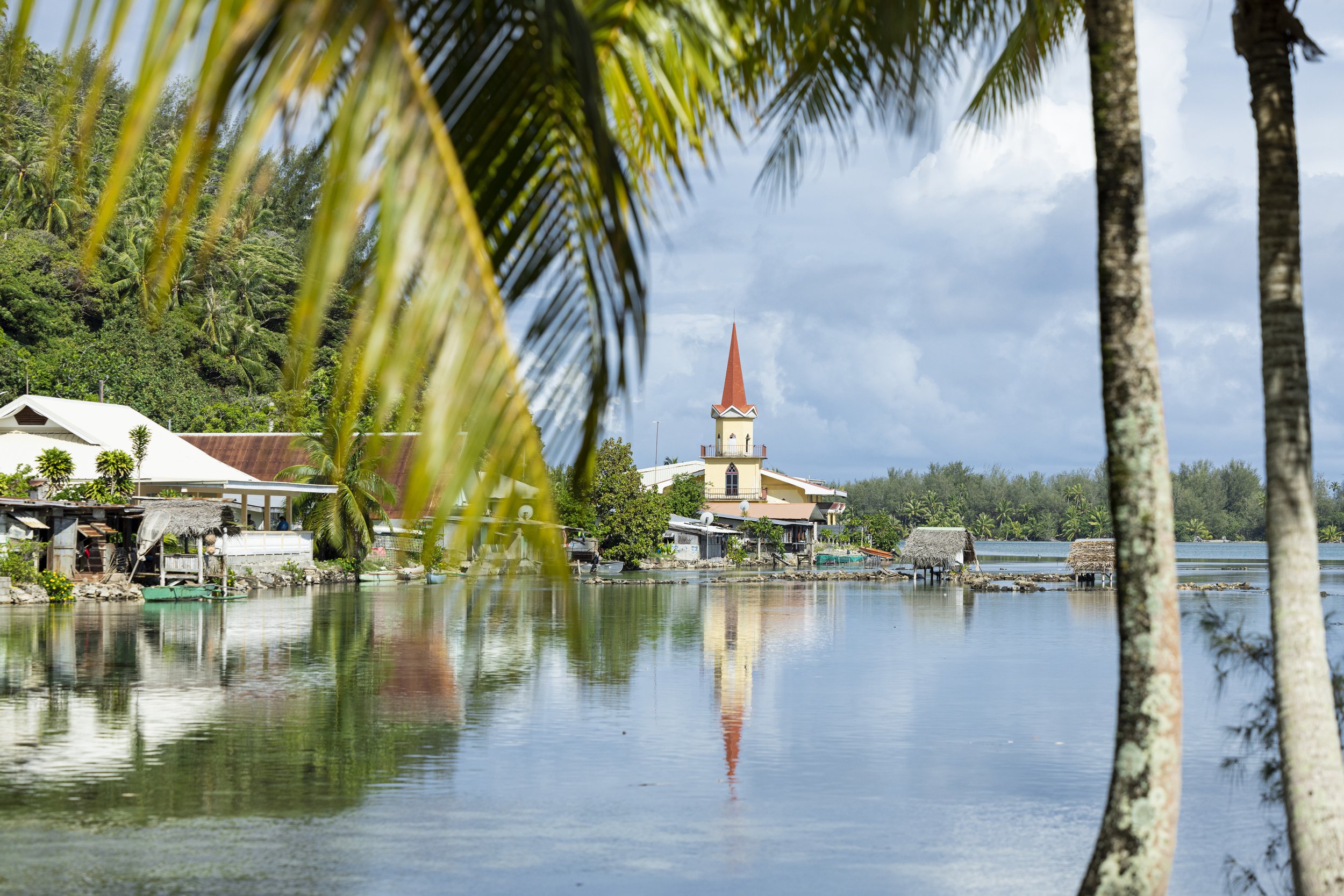 Vue d'une petite ville au bord de l'eau avec une église à clocher pointu, entourée de huttes, de maisons et de palmiers, sous un ciel partiellement nuageux.