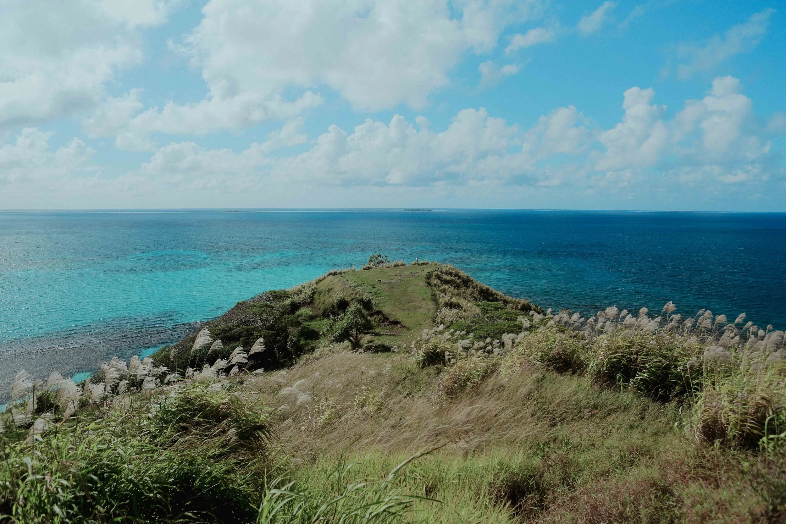 Paysage côtier avec une colline recouverte d'herbe et de végétation, face à une mer d'eau turquoise et profonde, sous un ciel partiellement nuageux.