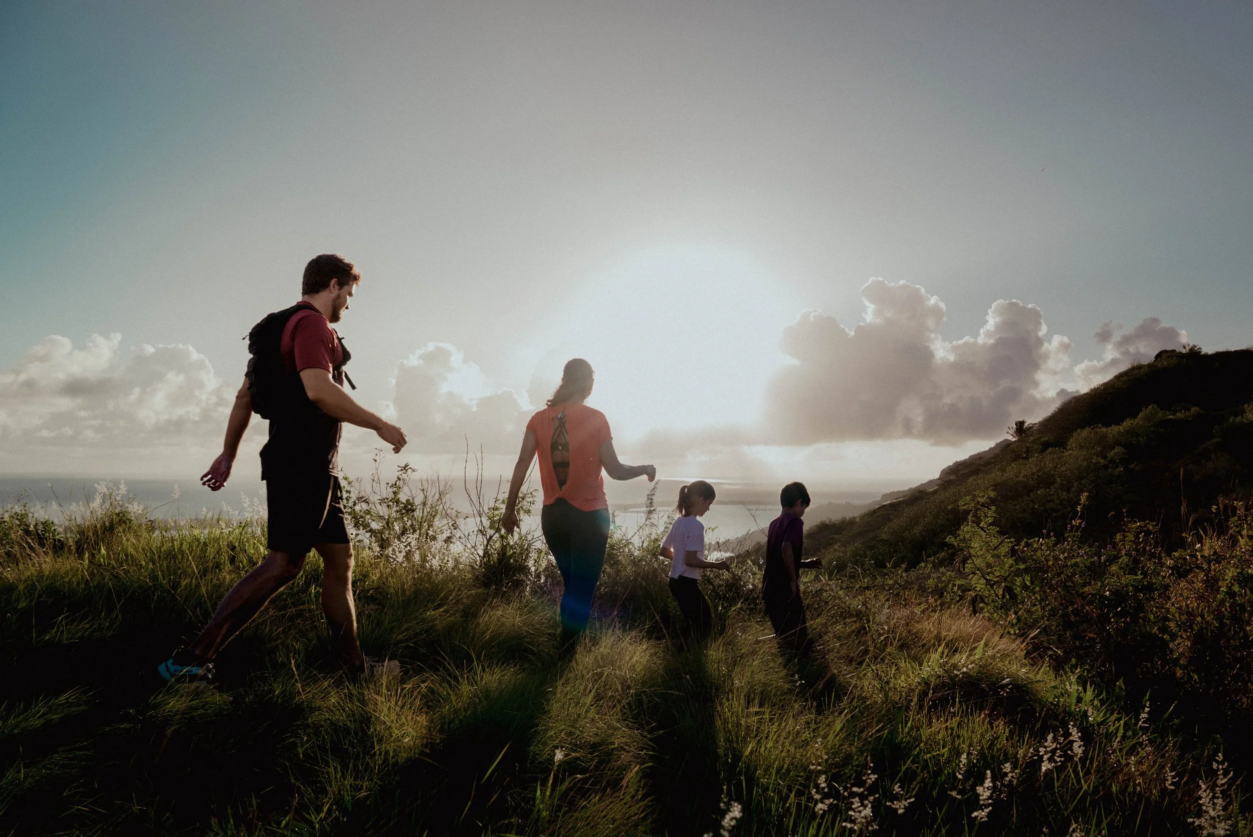 Une famille de cinq personnes faisant une randonnée dans un paysage naturel au coucher du soleil, dans des collines verdoyantes.