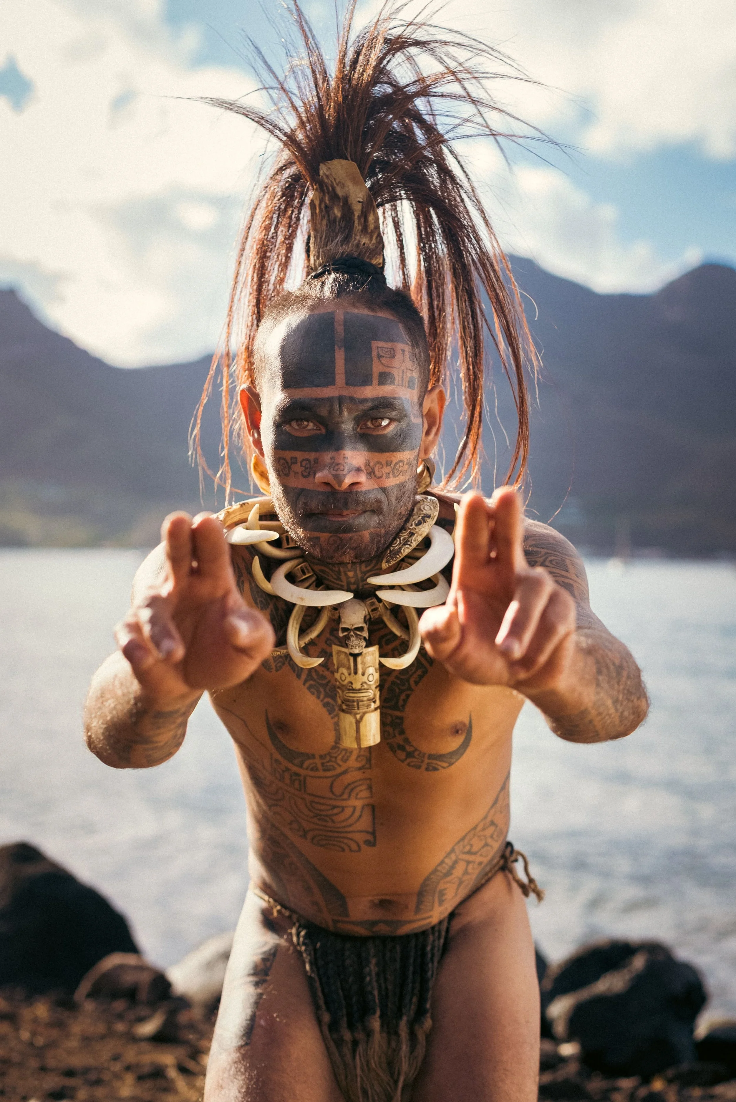 Homme moko nainé avec des tatouages et un collier en dents, pointant vers la caméra, près d'un lac avec montagnes en arrière-plan.