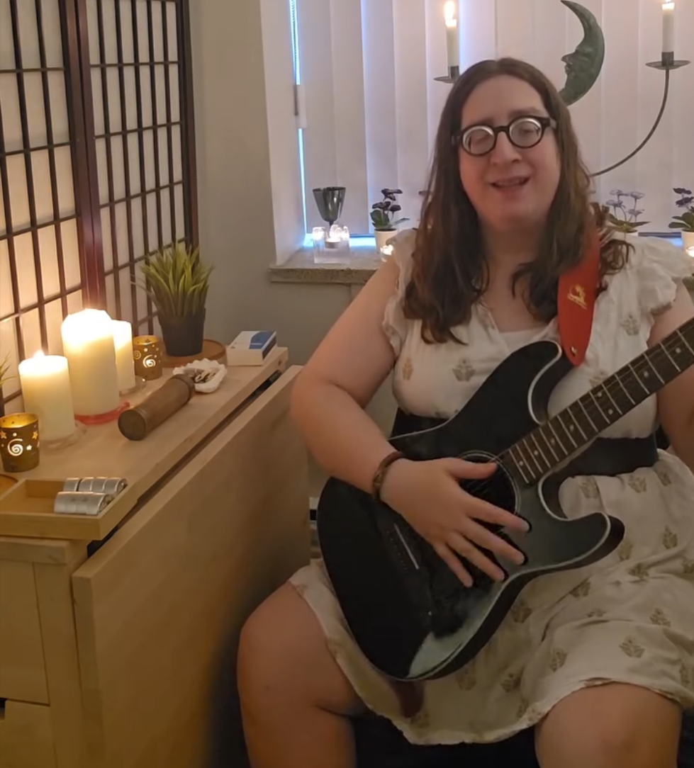 Aerissa in a white dress, with a black acoustic guitar surrounded by candles, wood, and plants