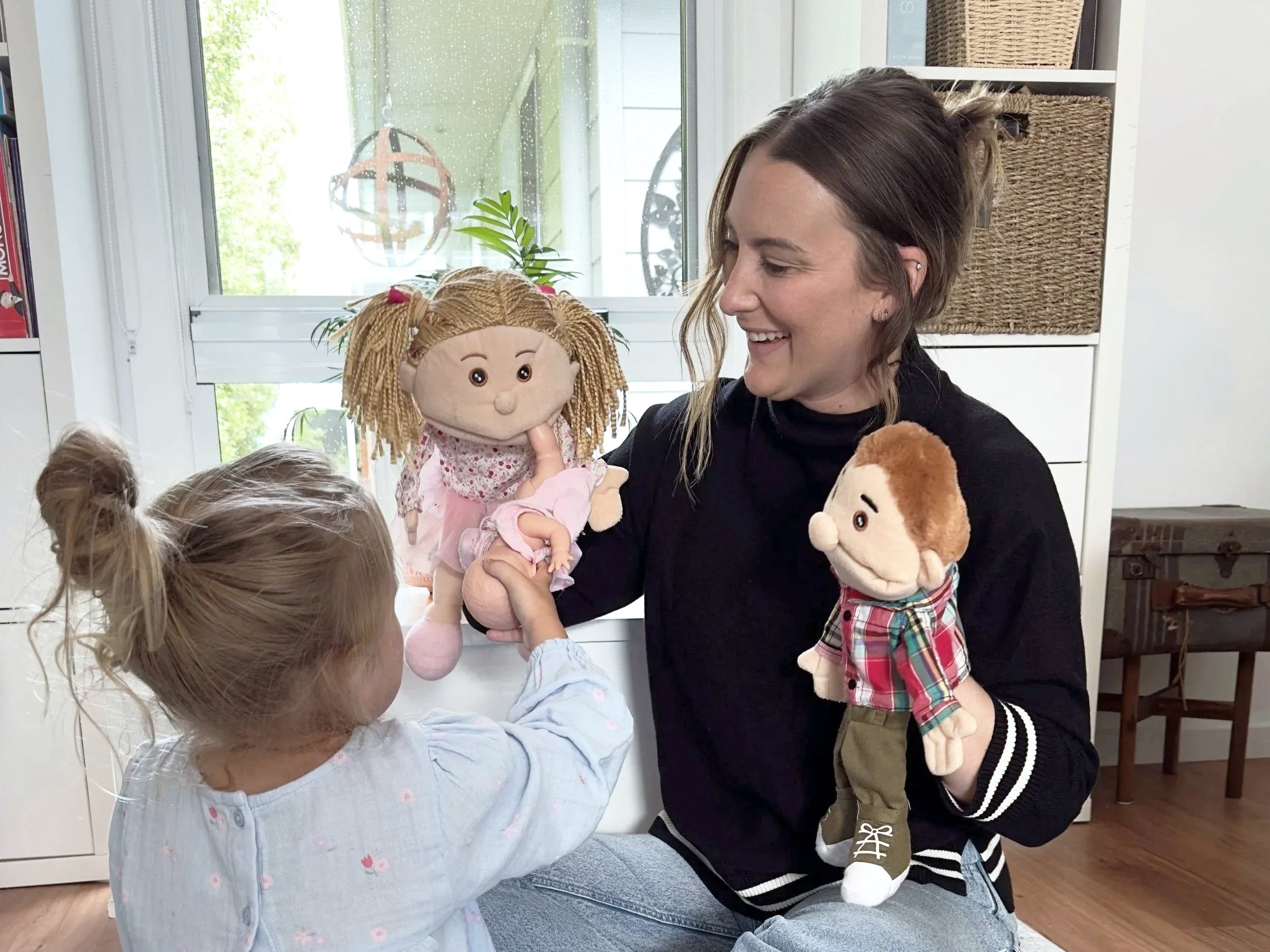 Various plush toys including a gray bunny, a white doll with a gold crown, a brown bear, and a green dinosaur with brown spikes, placed on and beside a light green and white striped box on a beige carpeted floor against a plain white wall.