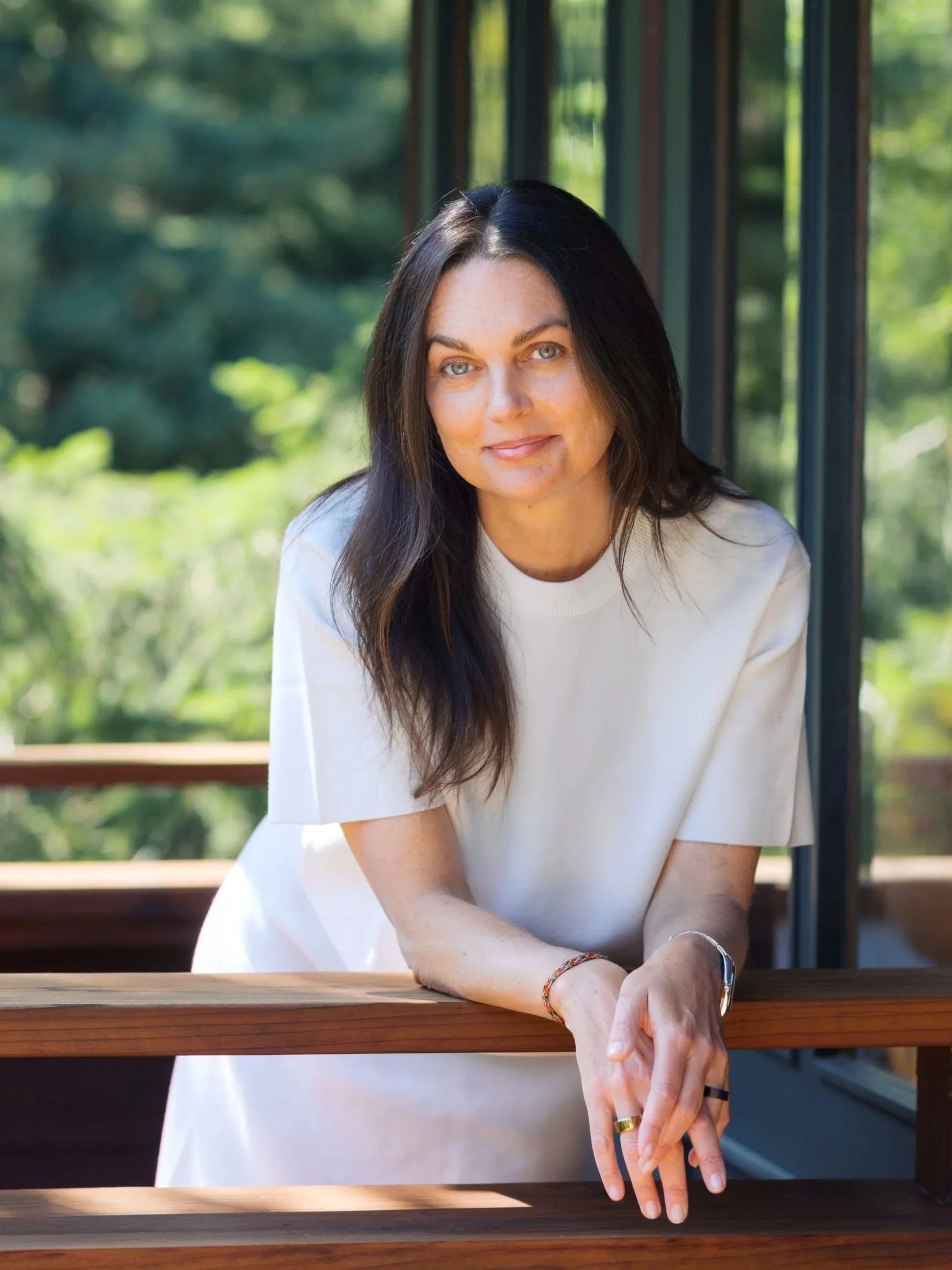 A woman with long dark hair and light skin leaning on a wooden railing, outdoors with greenery in the background.