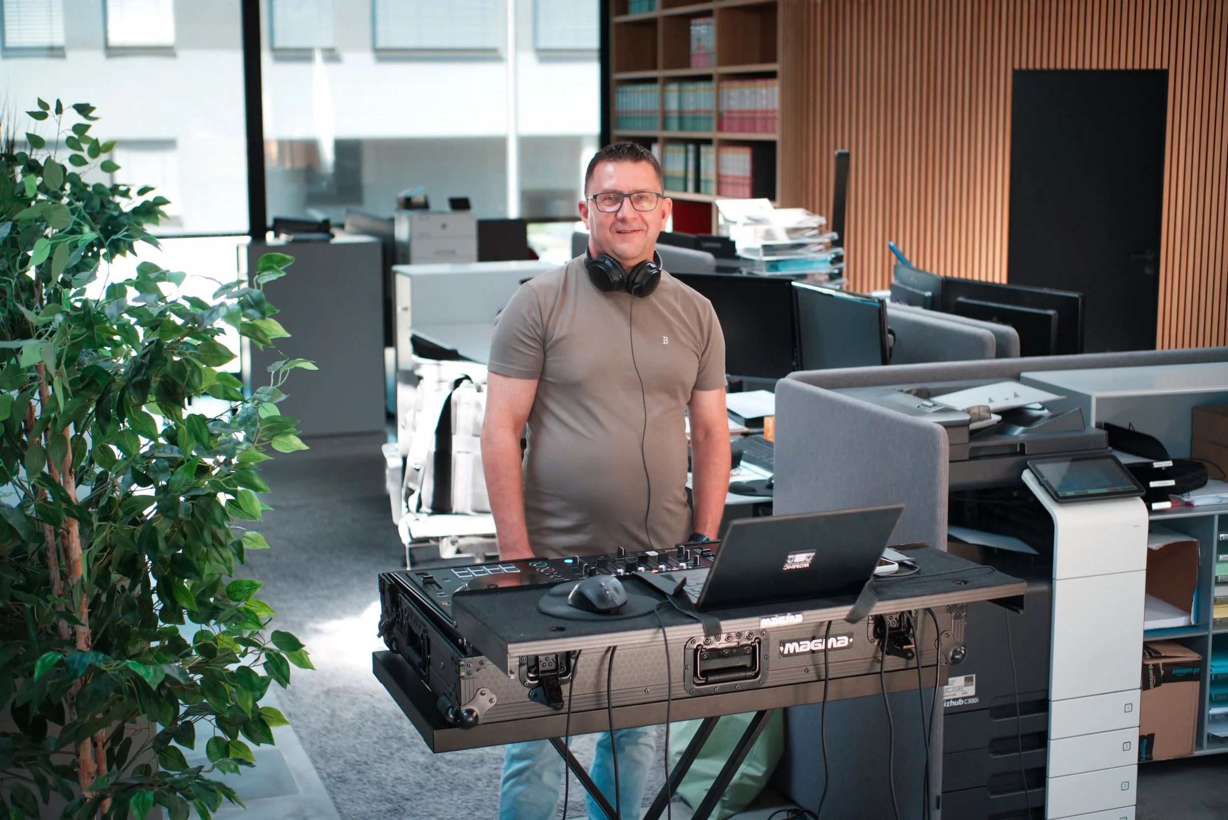 Ein Mann mit Brille, grau T-Shirt und Kopfhörern steht in einem modernen Büro hinter einem DJ-Controller auf einem Klavier. Im Hintergrund sind Schreibtische, Computer, Ordnerregale und große Fenster.