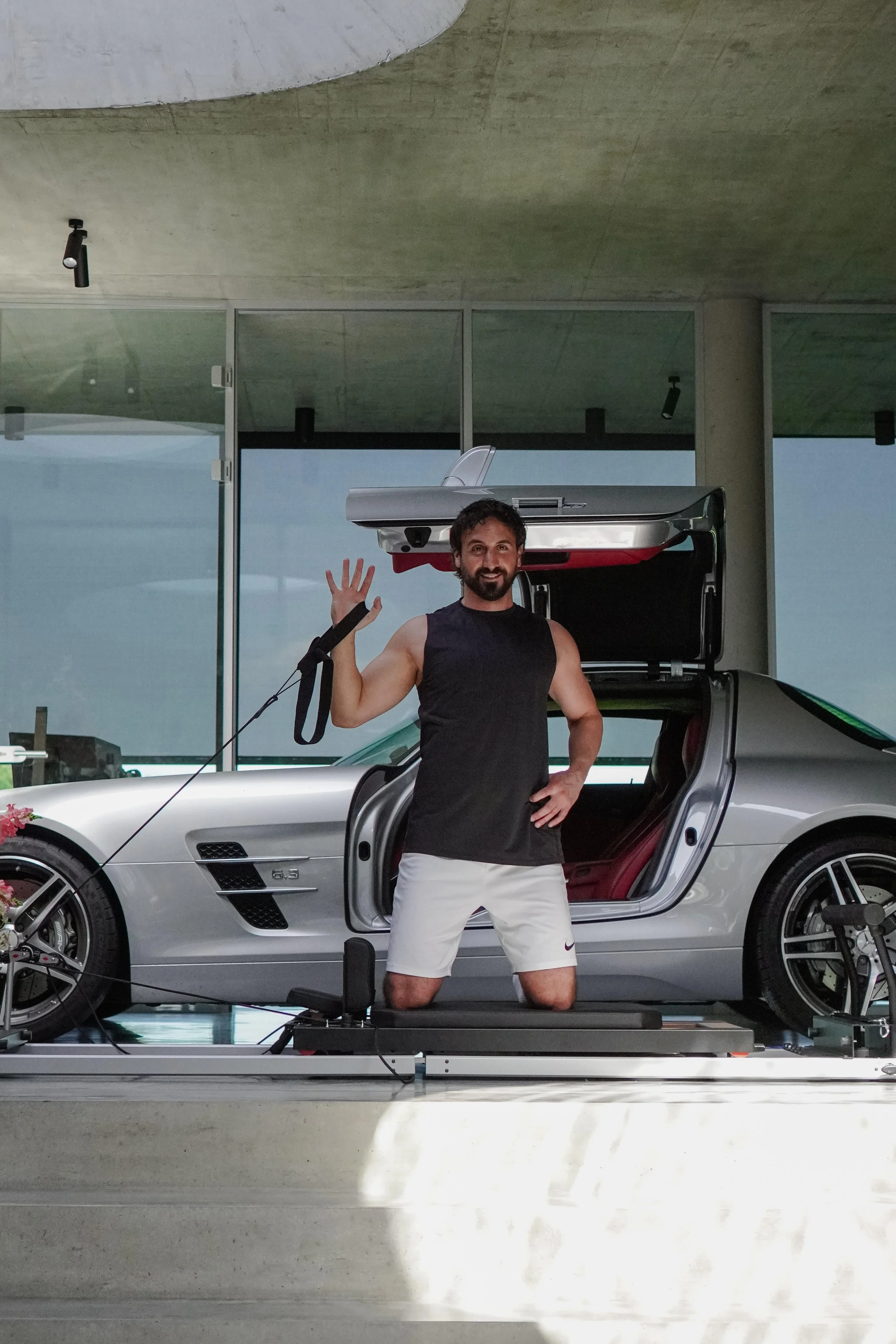 Ein Mann steht vor einem silbernen Sportwagen mit offenen Türen, in einem modernen Gebäude, und winkt zur Kamera.