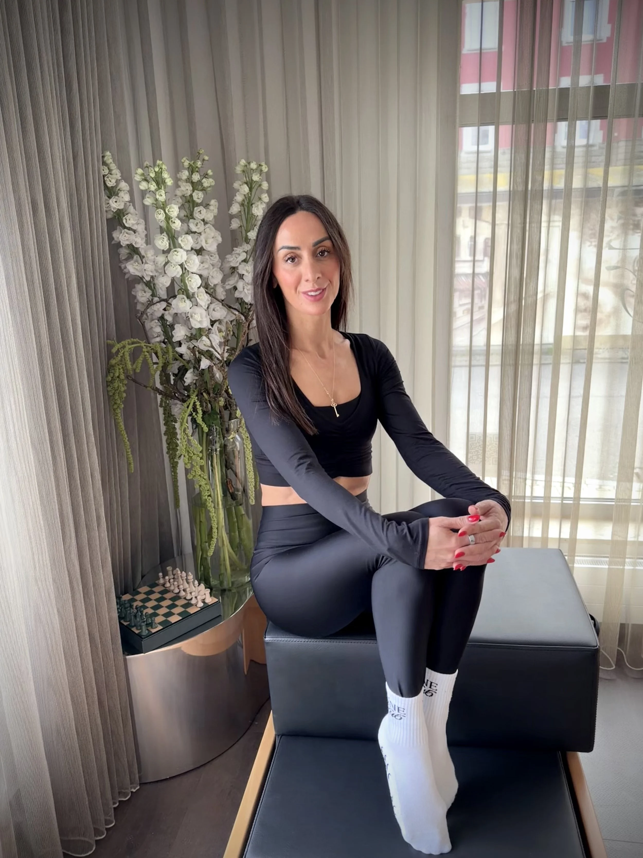 Eine Frau in schwarzer Sportkleidung, die auf einem schwarzen Stuhl in einem Raum mit großen Fenstern sitzt, neben einer Vase mit weißen Blumen und einem Schachspiel im Hintergrund.