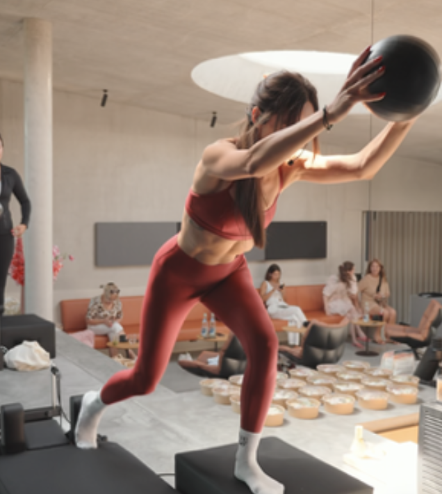 Frau beim Balance-Training mit Medizinball in einem Fitnessstudio, im Hintergrund sitzen mehrere Menschen auf einem Sofa und saufen Getränke.