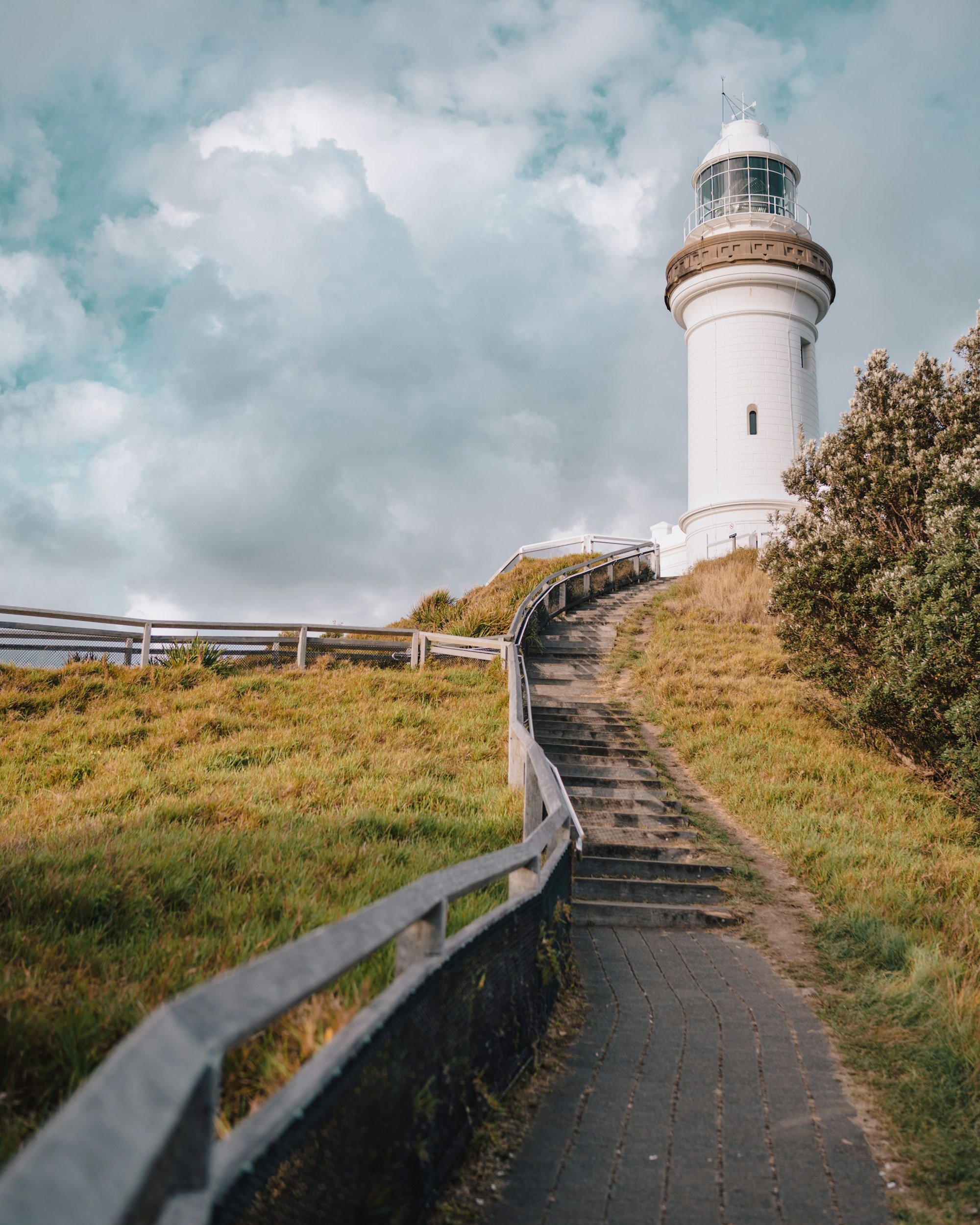 A white lighthouse on a hill with a winding wooden staircase and a railing, surrounded by grassy terrain and a bush, under a cloudy sky.