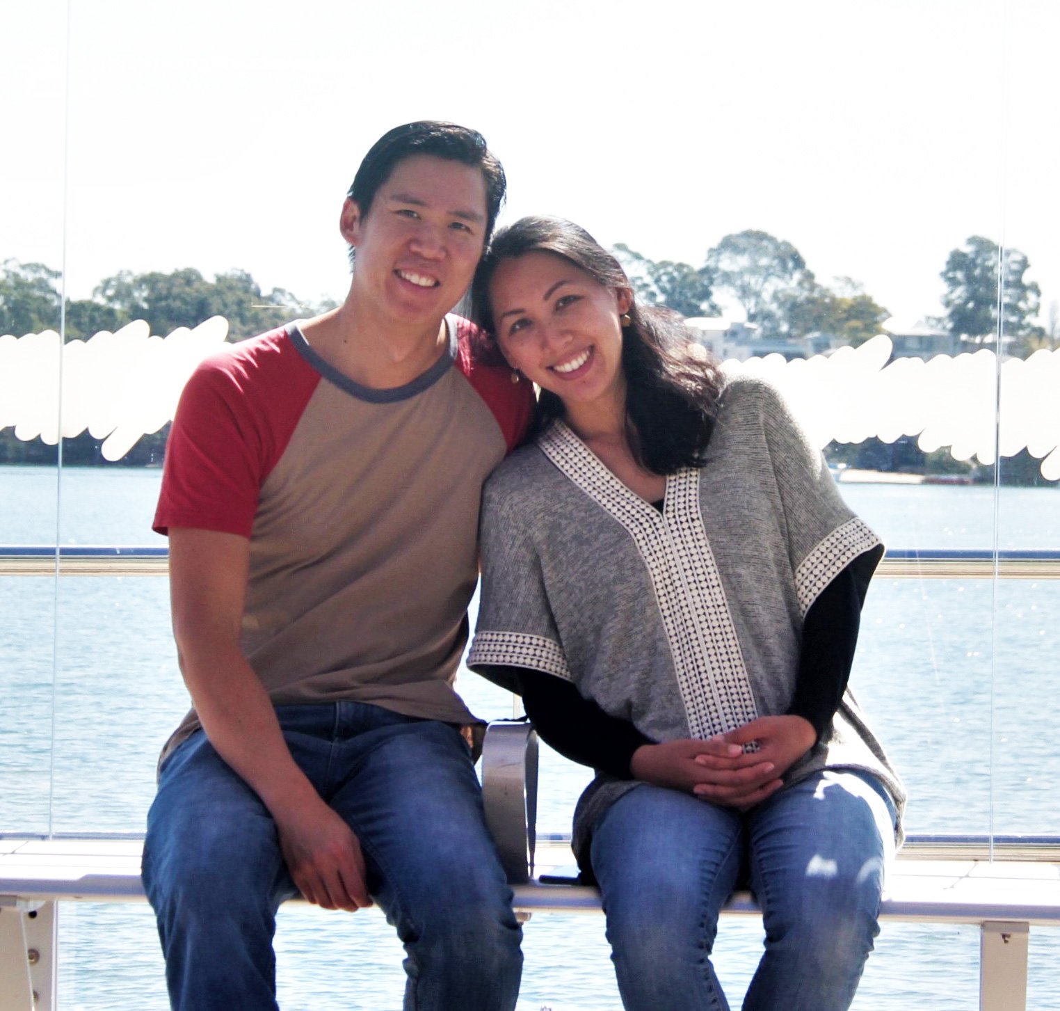 A smiling young man and woman sitting close together on a bench near a body of water, with trees and buildings in the background, during daytime.