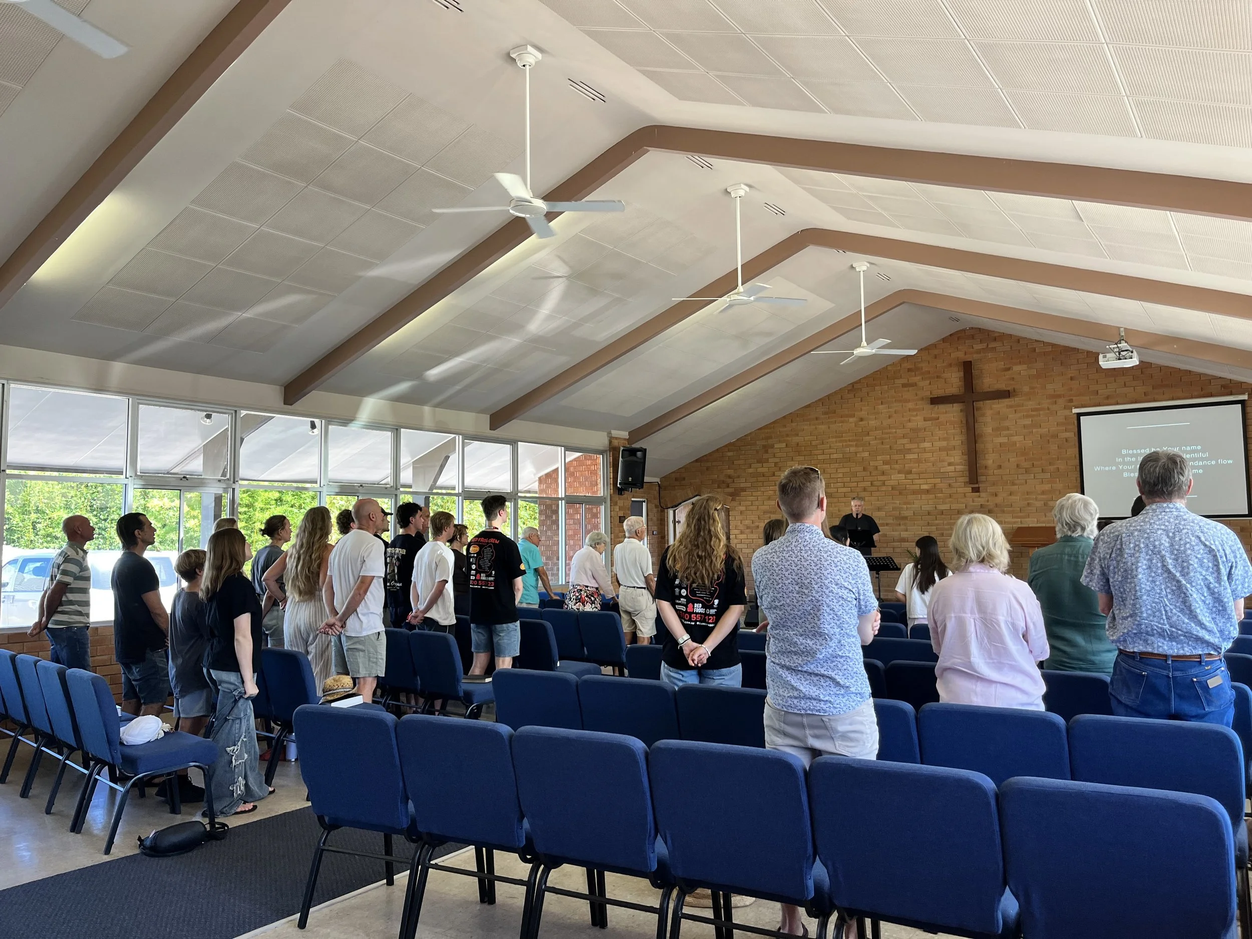 People standing in a church during a service, facing the front where a pastor is speaking, with a large cross on the brick wall behind him and a projector screen displaying song lyrics.