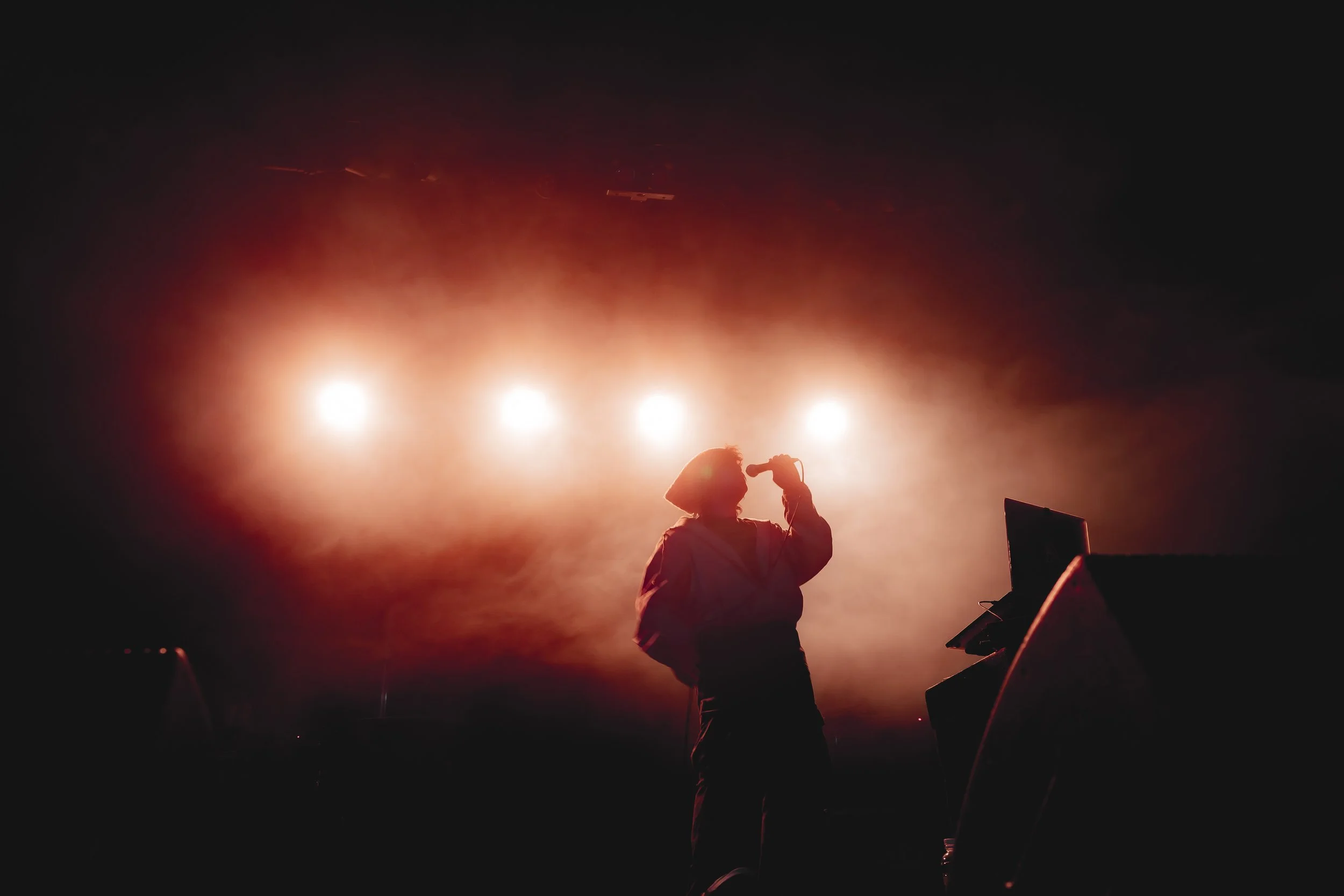 A singer performs on stage silhouetted against bright stage lights, with a microphone in hand and a laptop nearby.