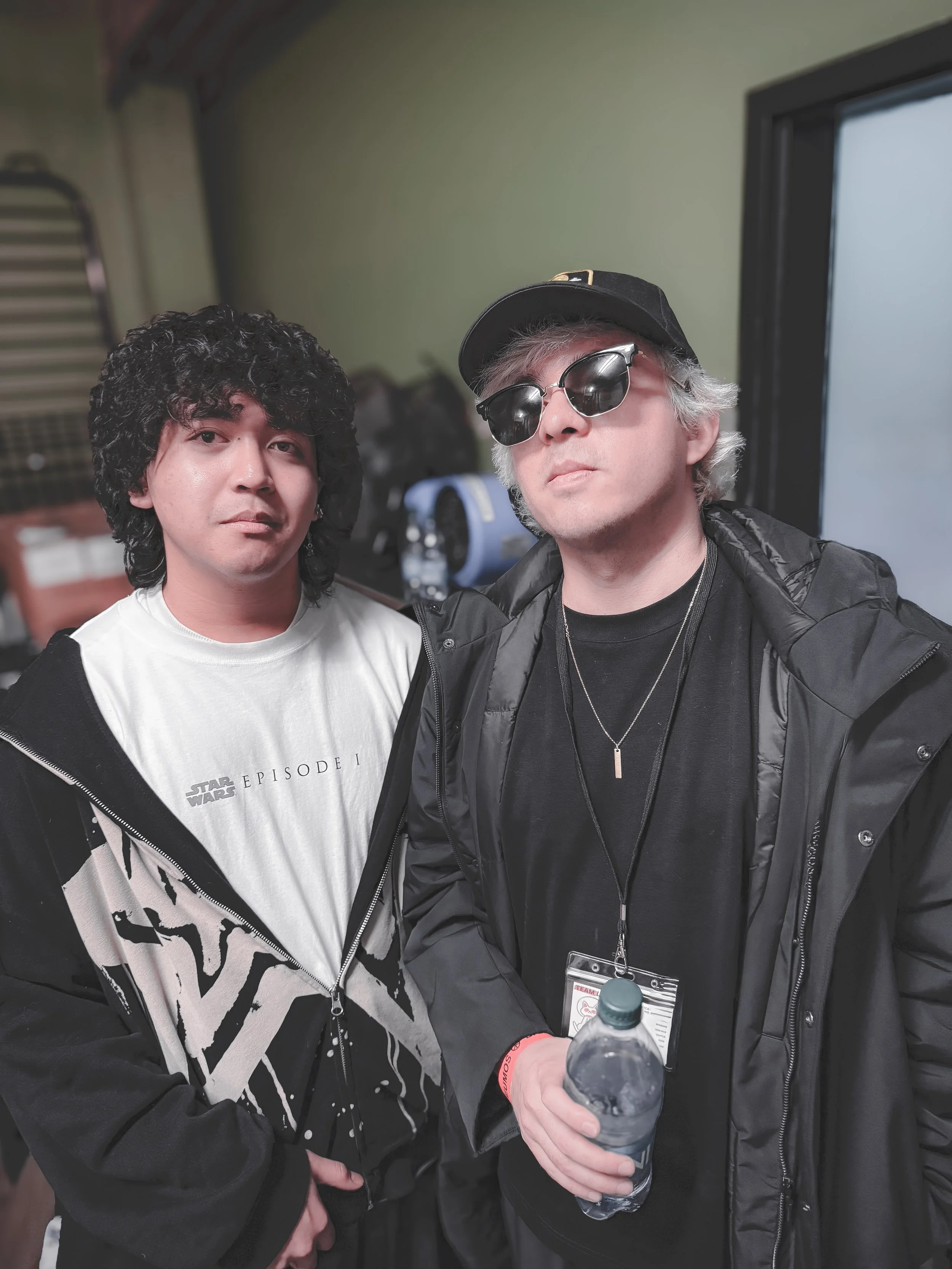Two men posing for a photo indoors. The man on the left has curly dark hair and is wearing a white t-shirt with 'Star Wars Episode I' printed on it, and a black and white jacket. The man on the right has silver hair, wears sunglasses, a black cap, bl