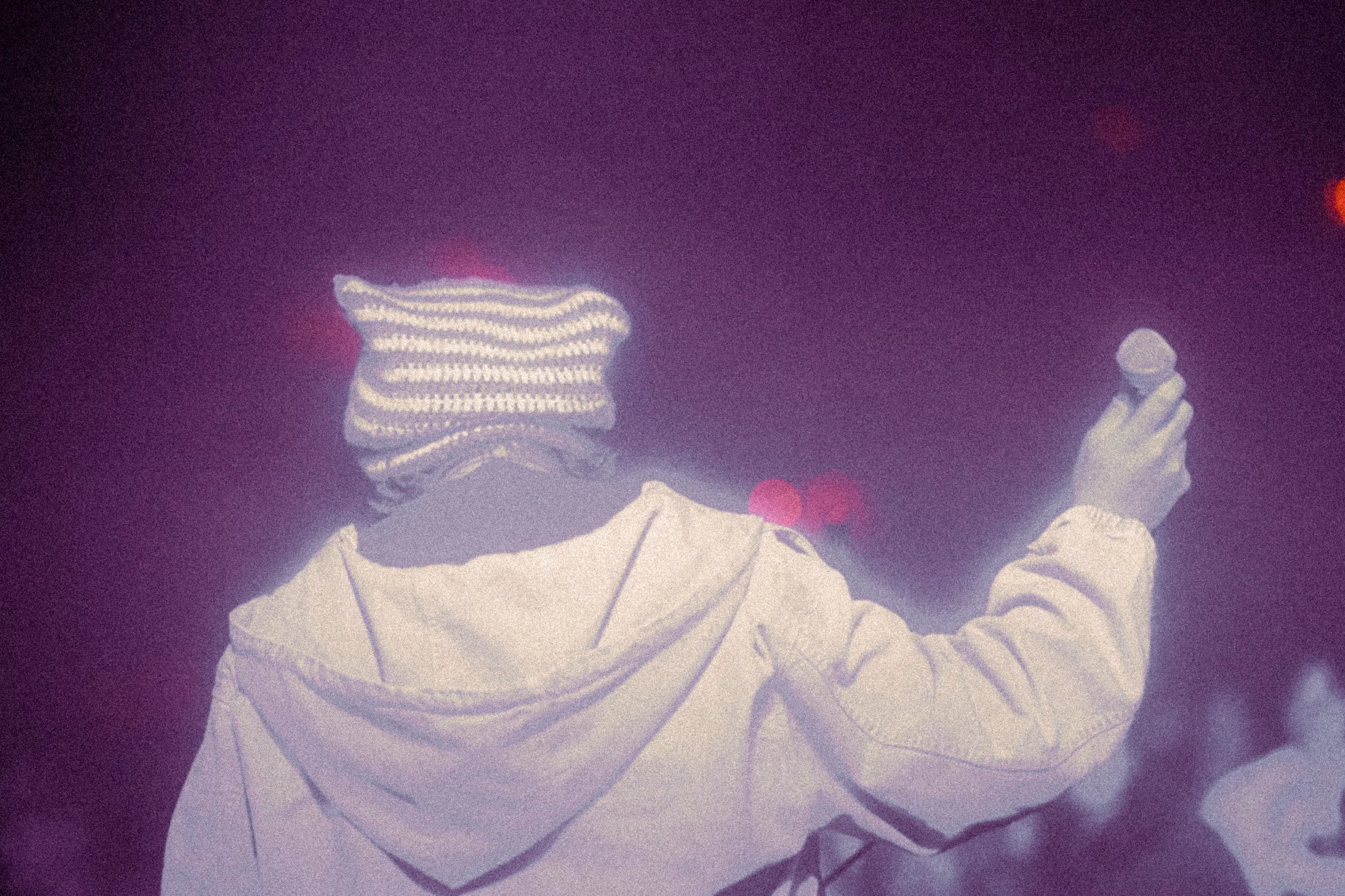 The singer "LCN!" in a white jacket holding a phone, with a striped hoodie underneath, in a dark environment.