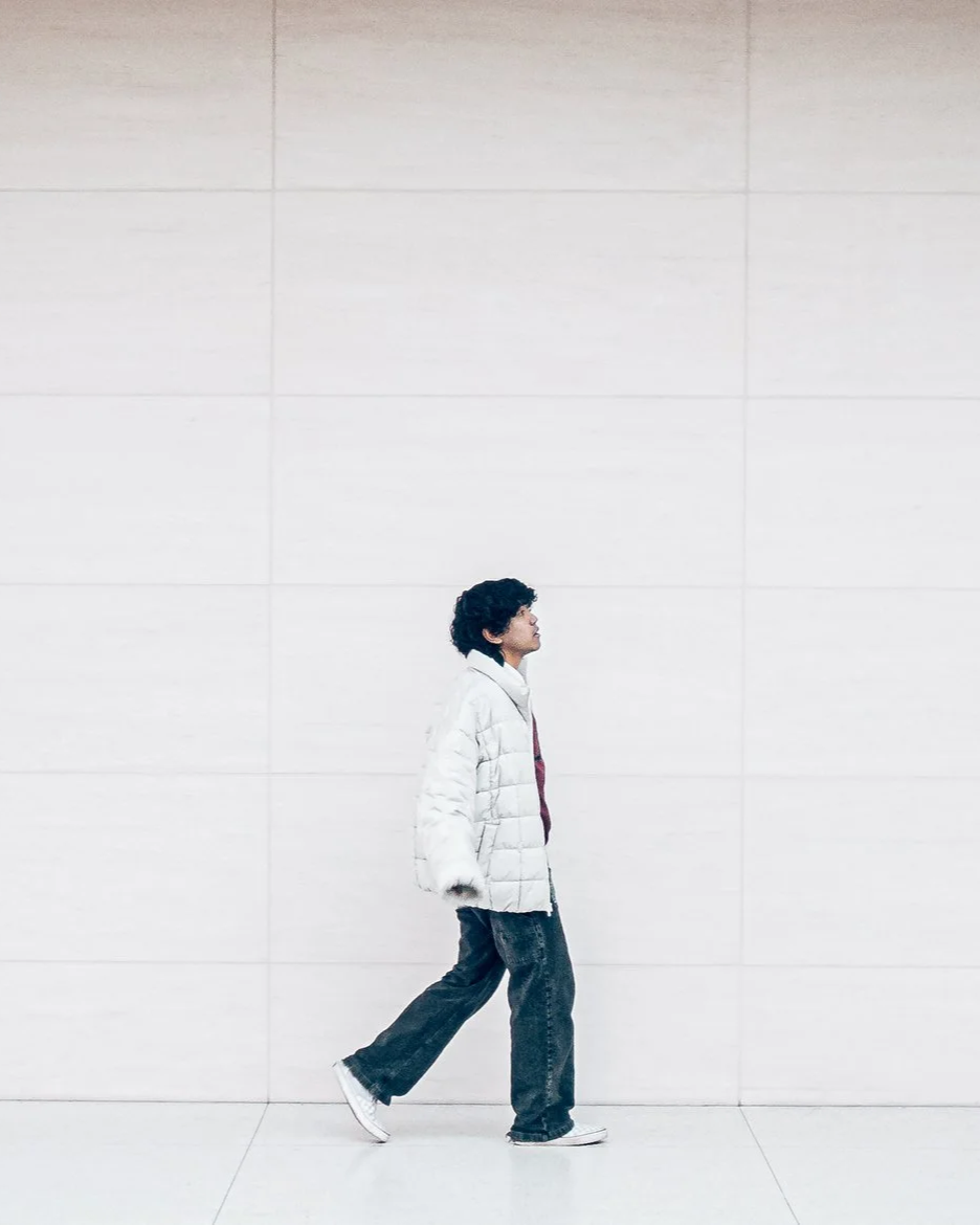 Young man walking indoors against a plain white tiled wall, wearing a white puffer jacket, jeans, and sneakers.