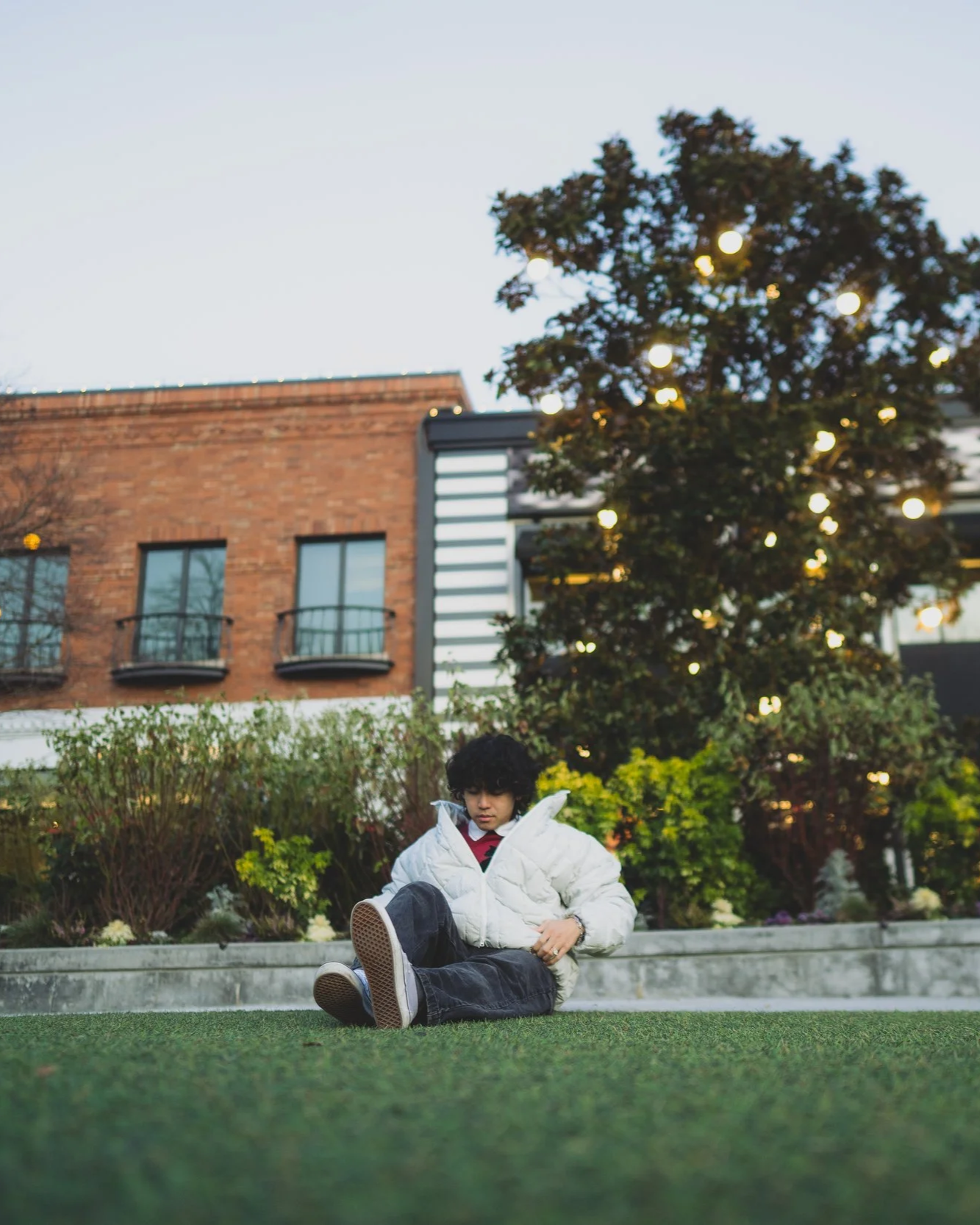 A person with curly black hair sitting on the grass in a park, wearing a white puffy jacket and black pants, in front of a landscaped garden with bushes and trees, with a brick building and a large tree decorated with string lights in the background.