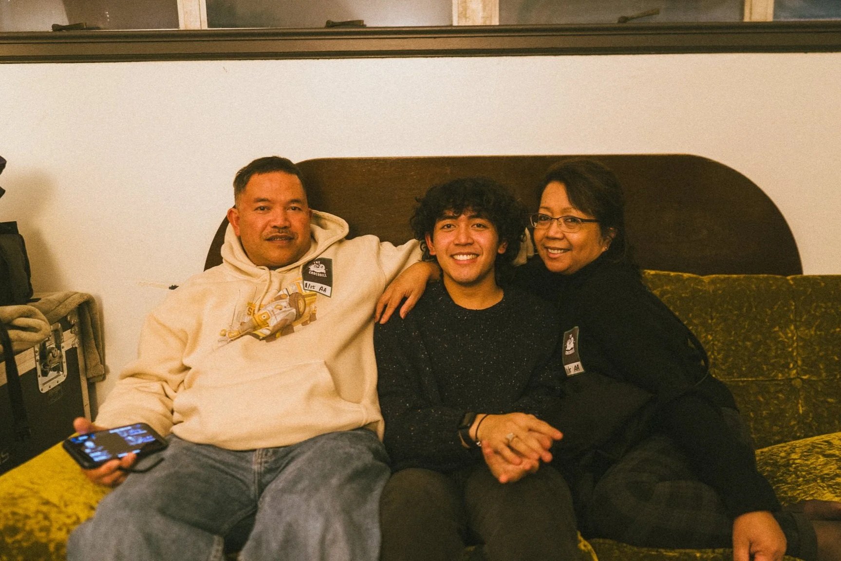 Three people sitting on a patterned yellow and black sofa, smiling at the camera. The person on the left is a man wearing a cream hoodie with a character design, holding a phone. The middle person is a young person with curly dark hair, wearing a bla