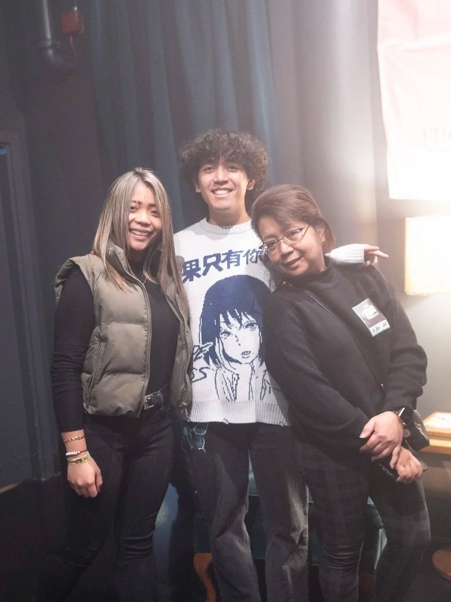 Three smiling people standing close together indoors, two women and one man, with dark curtains and a light source in the background.
