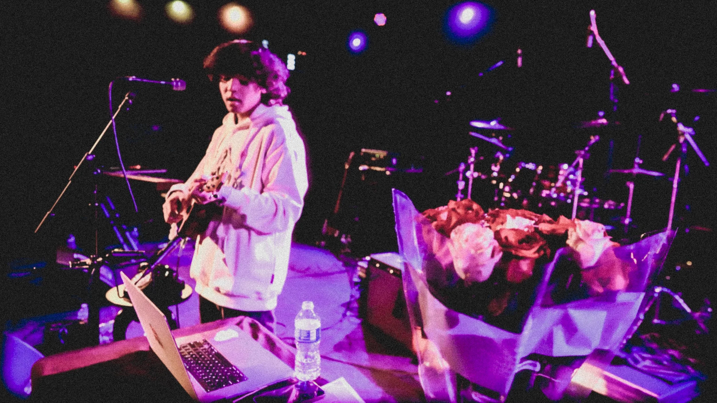 A person with curly hair wearing a light-colored hoodie is on stage with a microphone, a laptop, a water bottle, and a bouquet of flowers nearby, with musical instruments and stage lighting in the background.