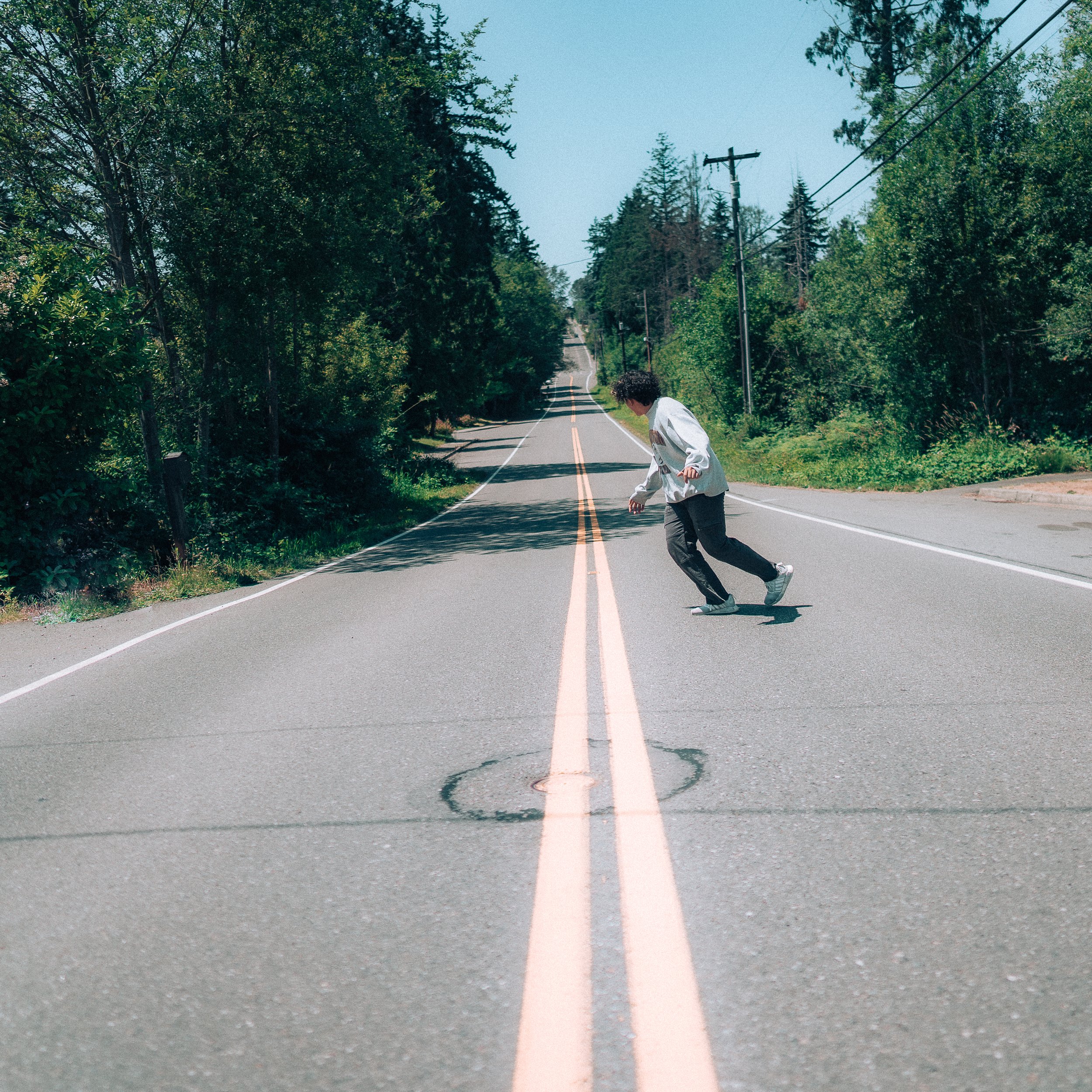 Person skateboarding in the middle of a quiet, empty rural road surrounded by green trees under a clear blue sky.