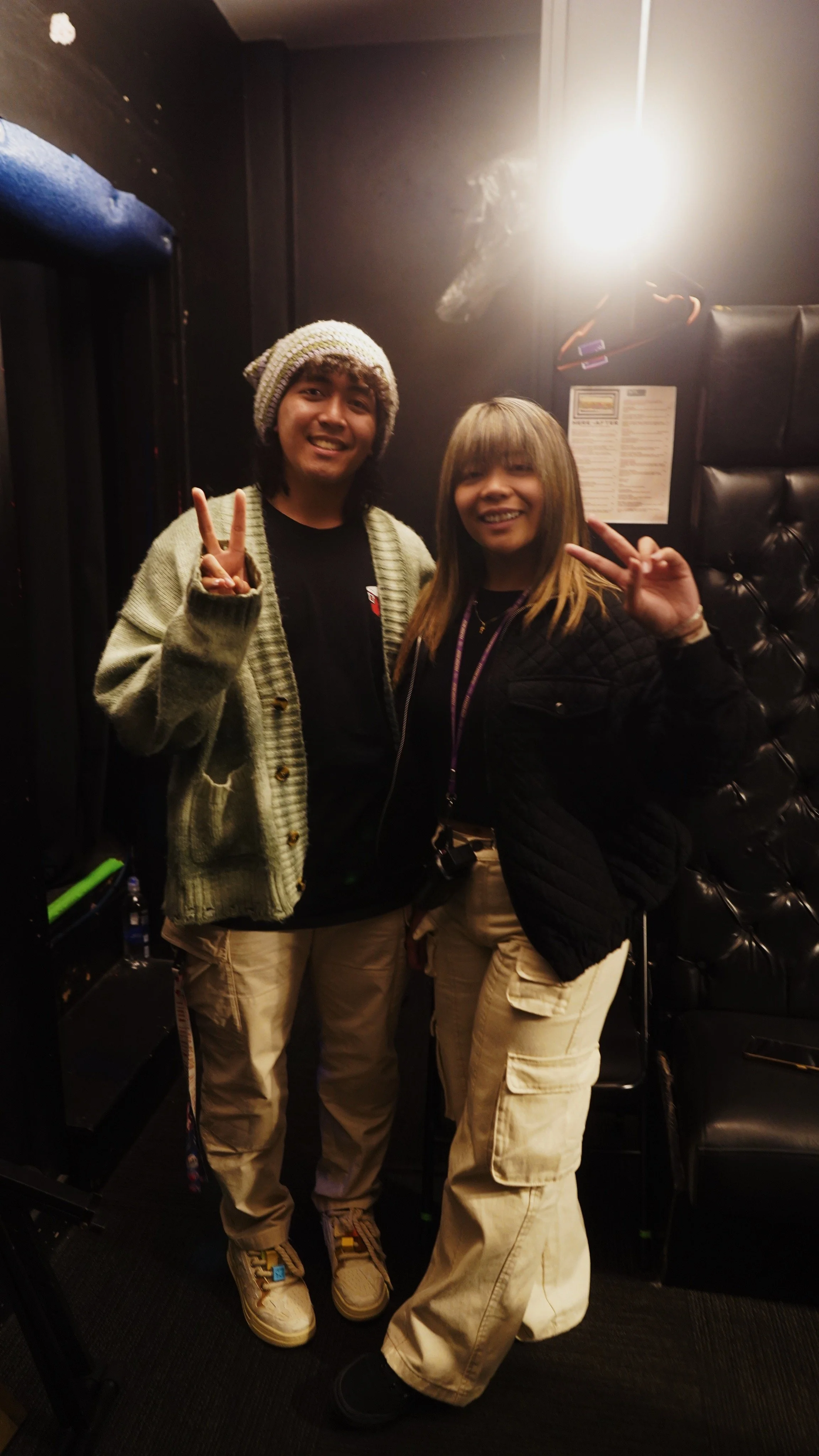 Two smiling people standing indoors, making peace signs with their hands. One is wearing a knit hat and cardigan, the other has straight hair and is dressed in a black jacket. The background includes black padded wall panels and a bright light source