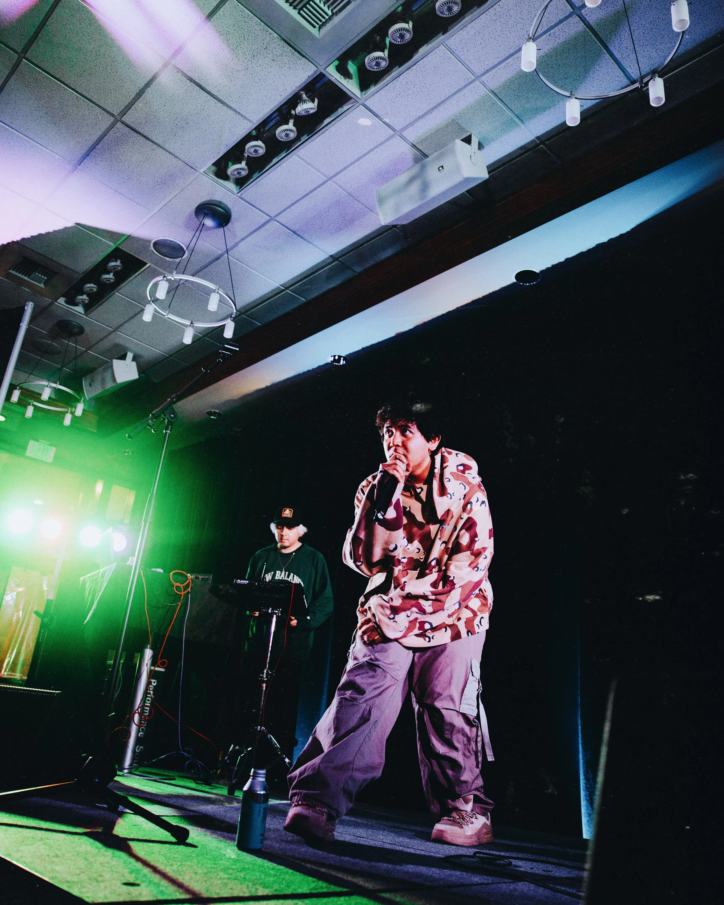 A performer holding a microphone and singing on stage with a DJ behind him, colorful stage lights, and a dark background.