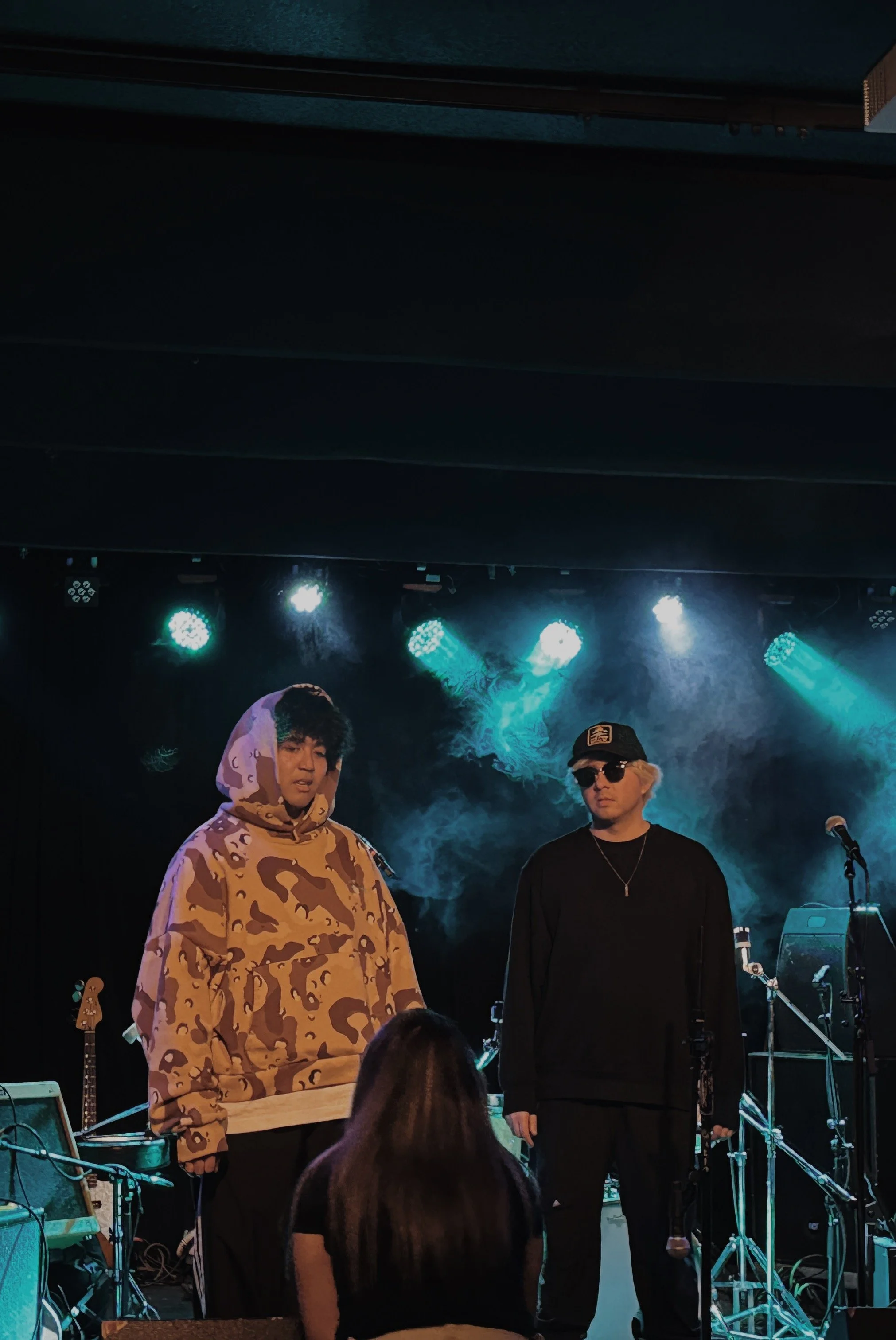 Two male performers on stage, one wearing a camo hoodie with the hood up, the other in black sunglasses and cap, with musical instruments and equipment around them, stage lights overhead, and an audience member in the foreground.